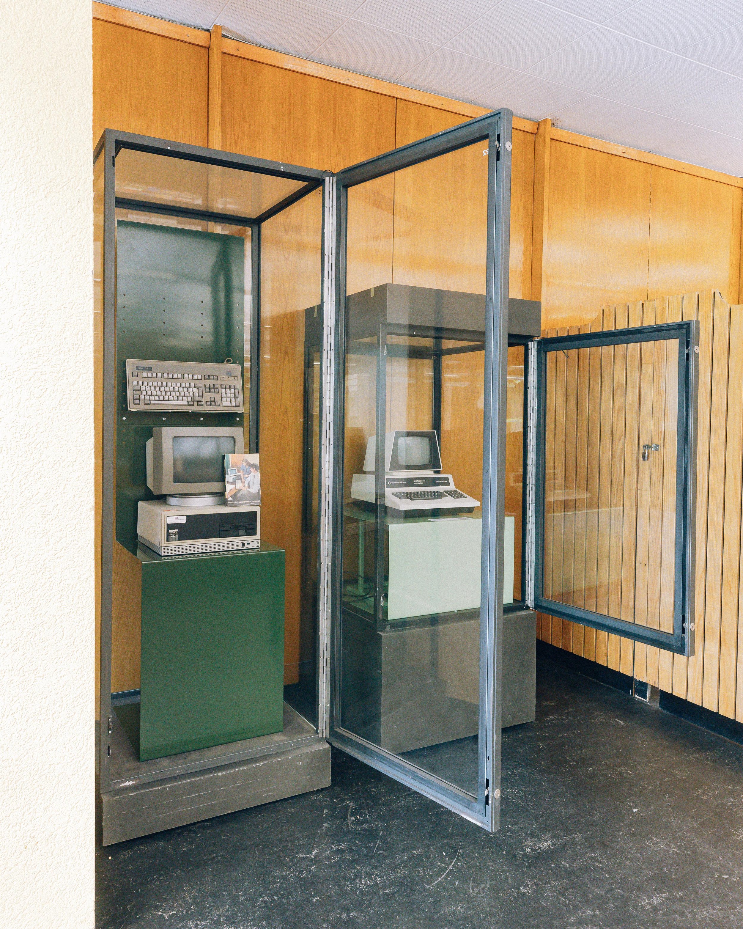 Une zone d'exposition contenant plusieurs anciens ordinateurs dans des vitrines en verre avec une porte ouverte, située dans une pièce avec des murs en bois et un plafond blanc.