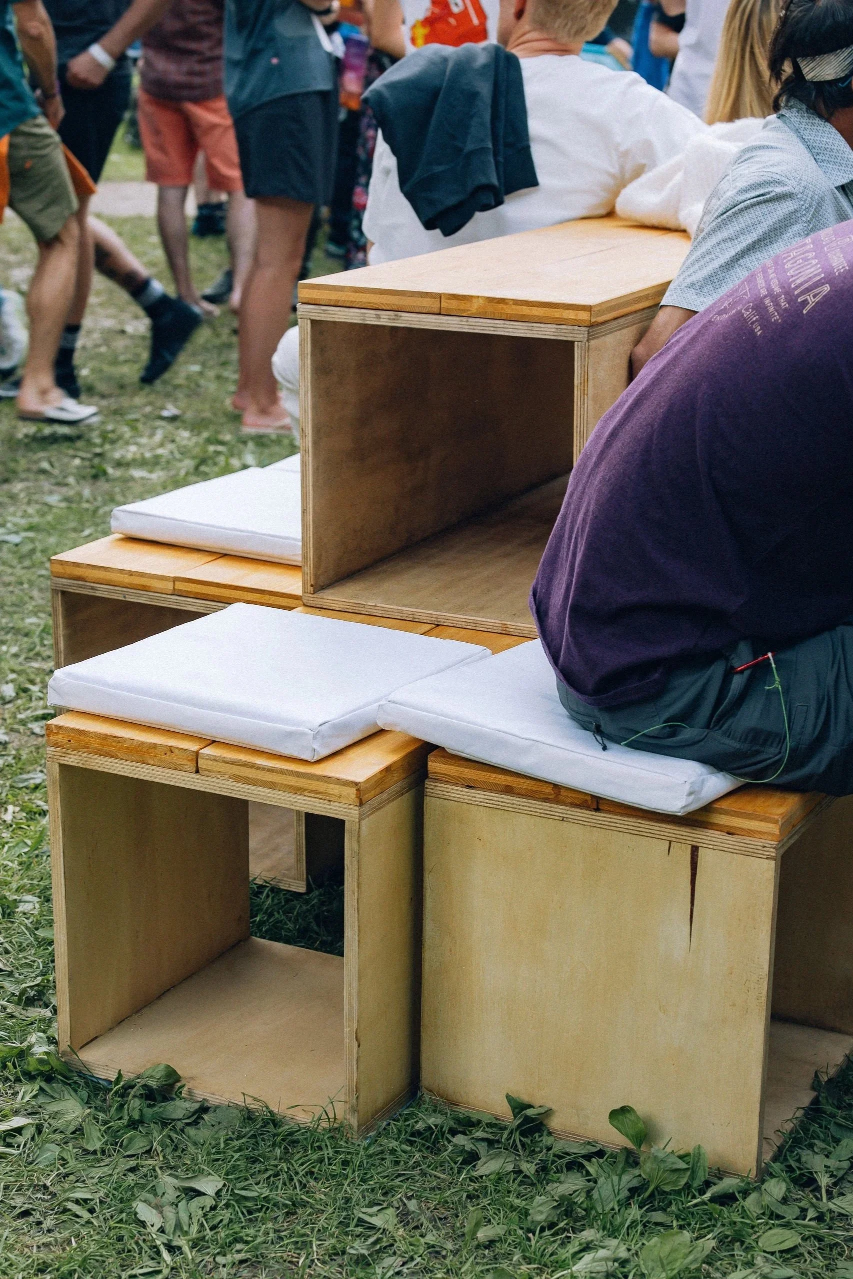 Des personnes s'asseyant sur des bancs en bois avec des coussins blancs lors d'un événement en plein air, entouré d'herbe coupée.