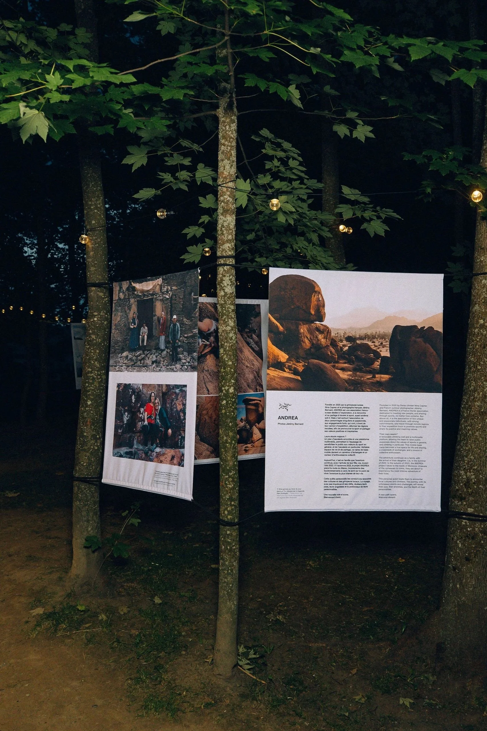 Exposition photographique suspendue dans un espace naturel, affichant plusieurs panneaux avec des images de paysages, personnes et textes. L'ensemble est accroché à des cordes entre deux arbres.