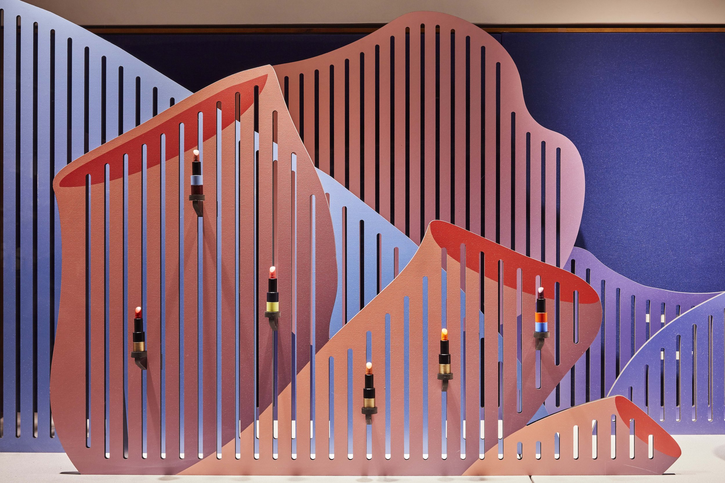 Une scénographie Hermès composée de différentes formes de coraux de couleurs rose, violet et bleu. La sculpture est percée de plusieurs bandes verticales etmet en lumière des rouges à lèvre grâce à un système scénographique modulable.