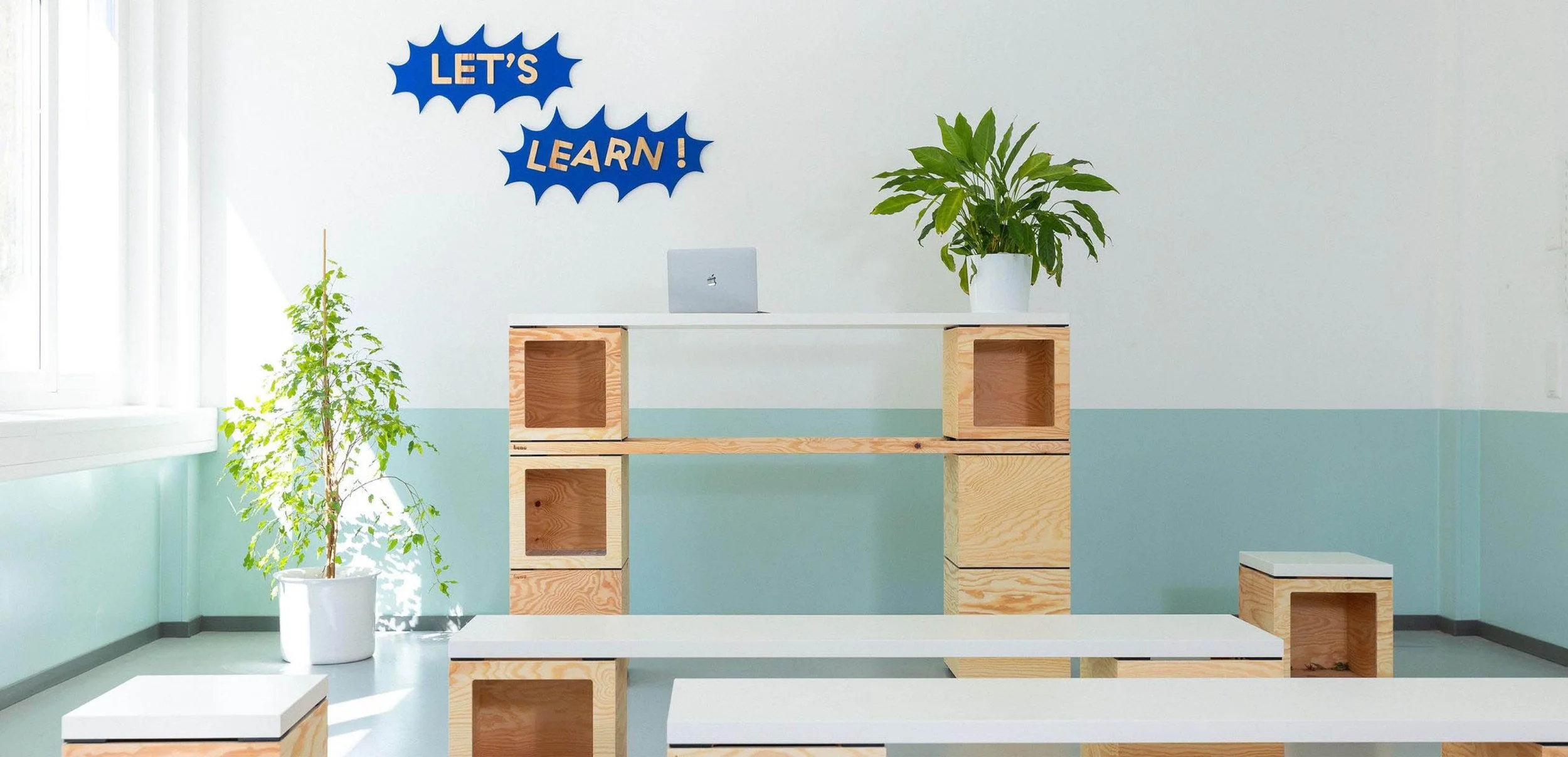 Une salle de workshop dans un coworking avec du mobilier modulable en bois clair, un bureau et étagères, des bancs,  deux plantes, un mur blanc avec une décoration en forme de nuages bleus avec des inscriptions en français « LET'S LEARN ! ».