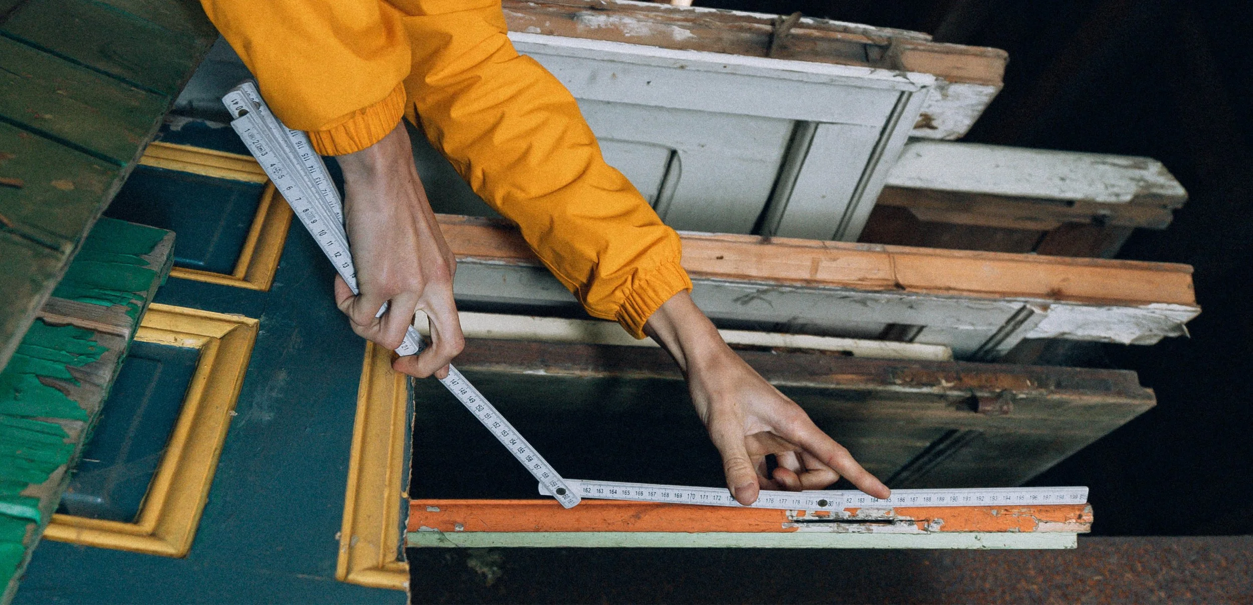 Une personne en veste orange mesure une pièce avec un mètre ruban dans un entrepôt de bois avec des planches empilées en arrière-plan.
