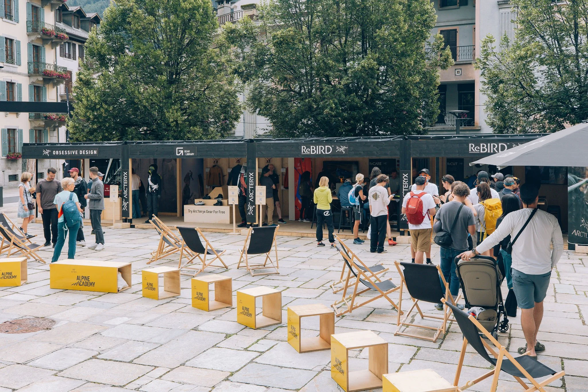 Une scène dans une place publique avec de nombreuses personnes, des chaises en bois discrètes, des stands de vente ou d'information, et des arbres verts en arrière-plan, dans une atmosphère de rassemblement ou d'événement.