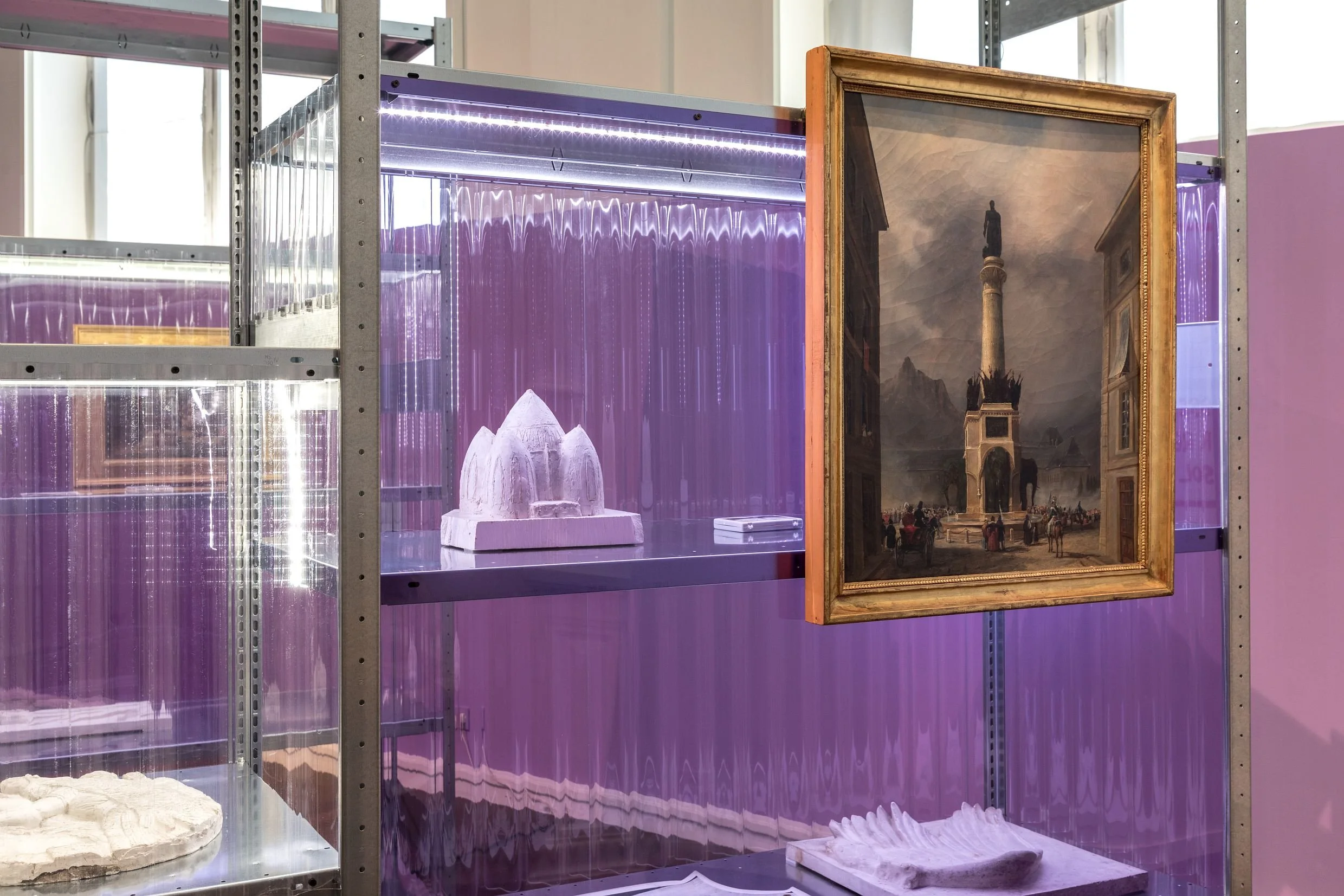Une pièce d'exposition avec des œuvres d'art, notamment une maquette en plâtre d'une structure architecturale, un tableau représentant une scène urbaine avec un monument, et des objets architecturaux en plâtre.