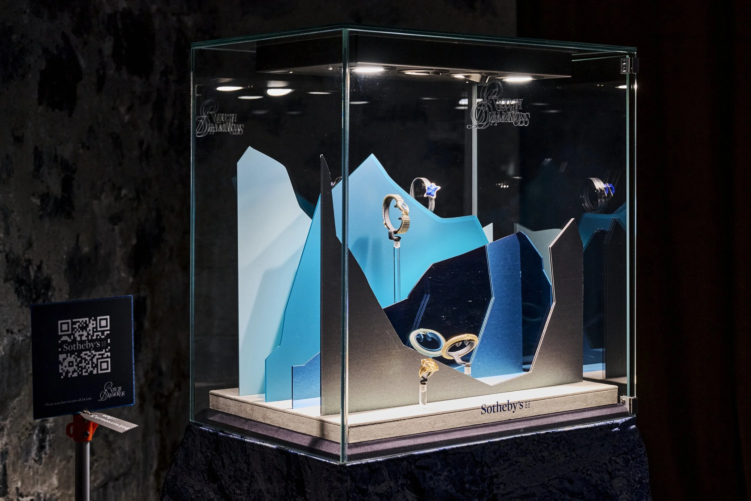 Une vitrine contenant plusieurs bijoux, notamment des bagues et des bracelets, exposés sur un fond bleu et blanc, sous un éclairage brillant. À gauche, un panneau avec un QR code et le logo de Sotheby's.