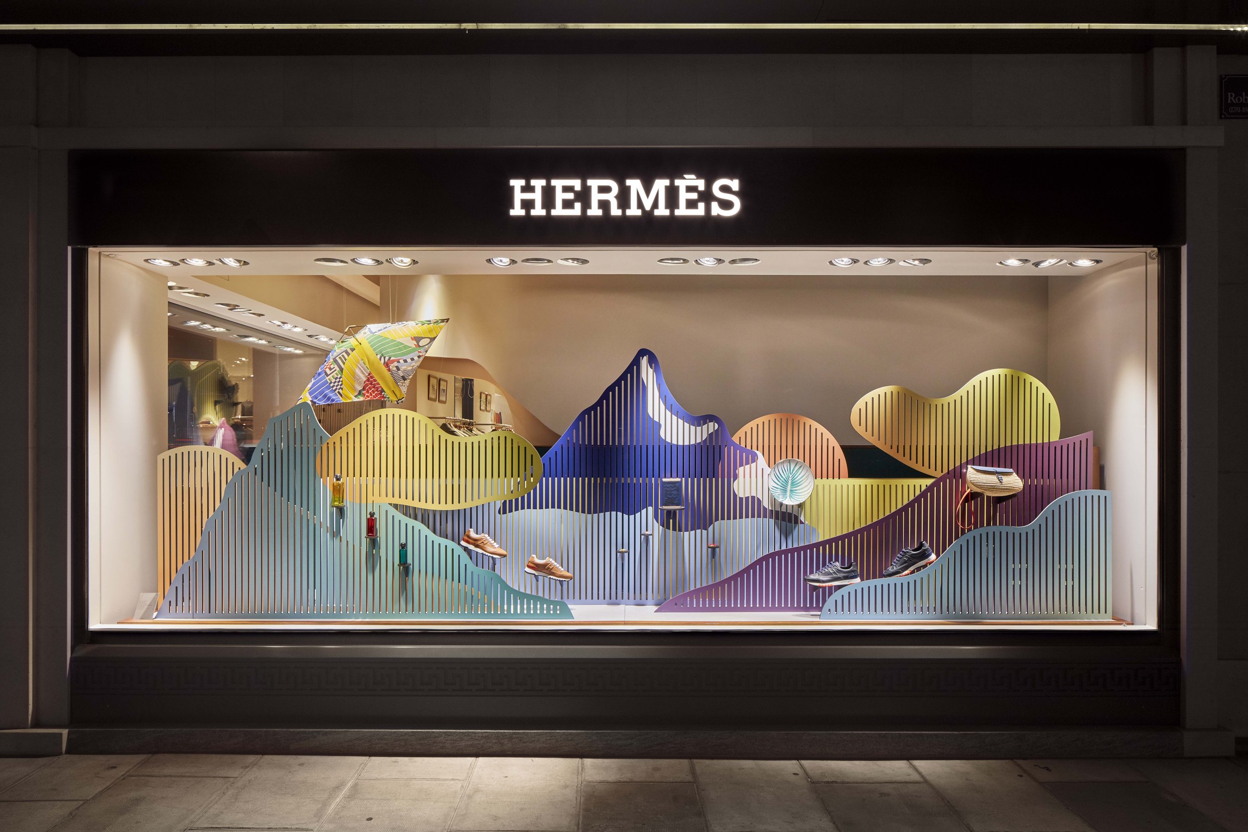 Vitrine d'une boutique Hermès avec scénographie colorée représentant des paysages montagneux en Suisse, mettant en valeur des objets Hermès tels que des parfums, un foulard, des chaussures, une assiette, un sac à main.