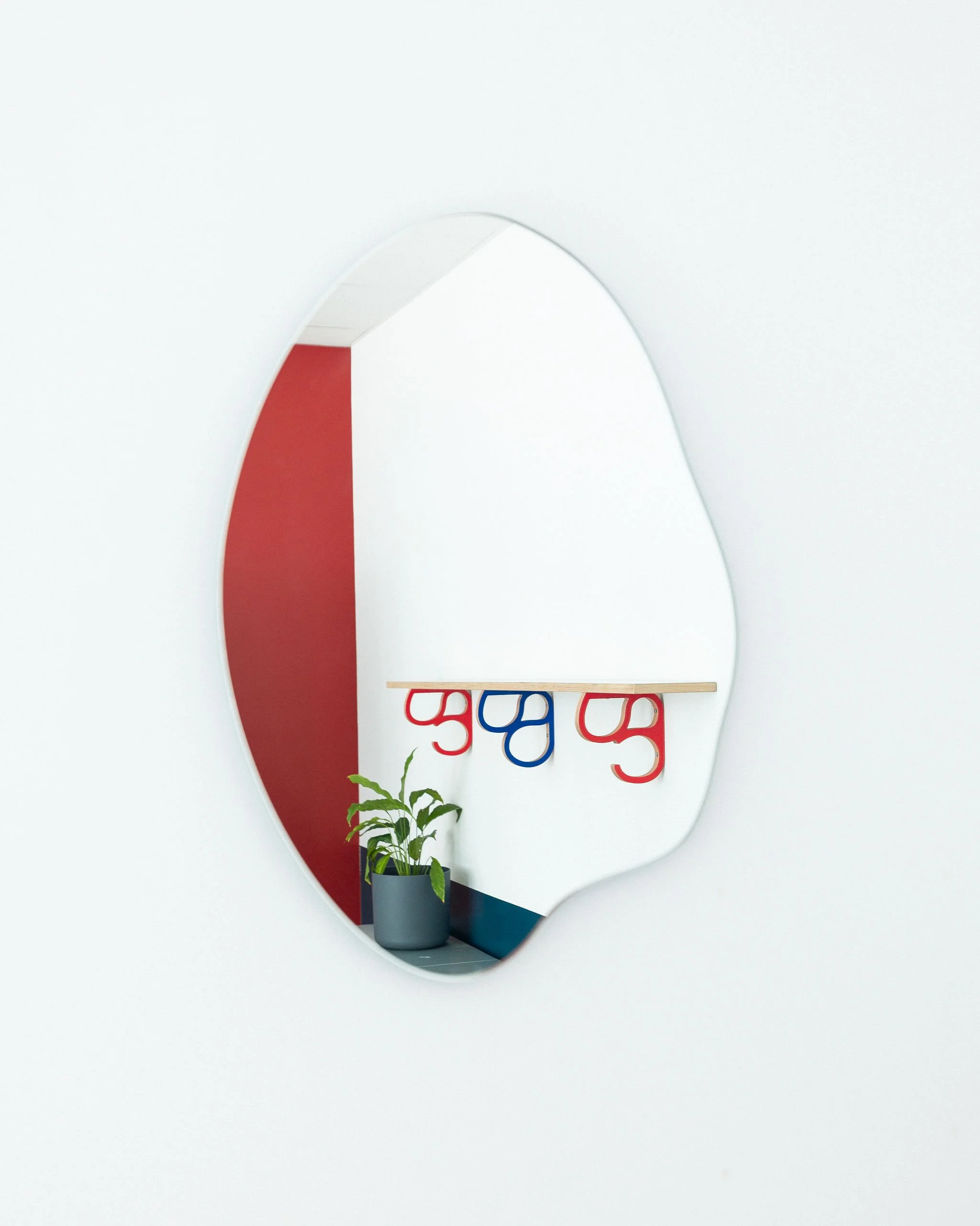 Un miroir mural de forme organique reflétant un mur rouge, une étagère avec des patères aux formes organiques et colorées et une plante.