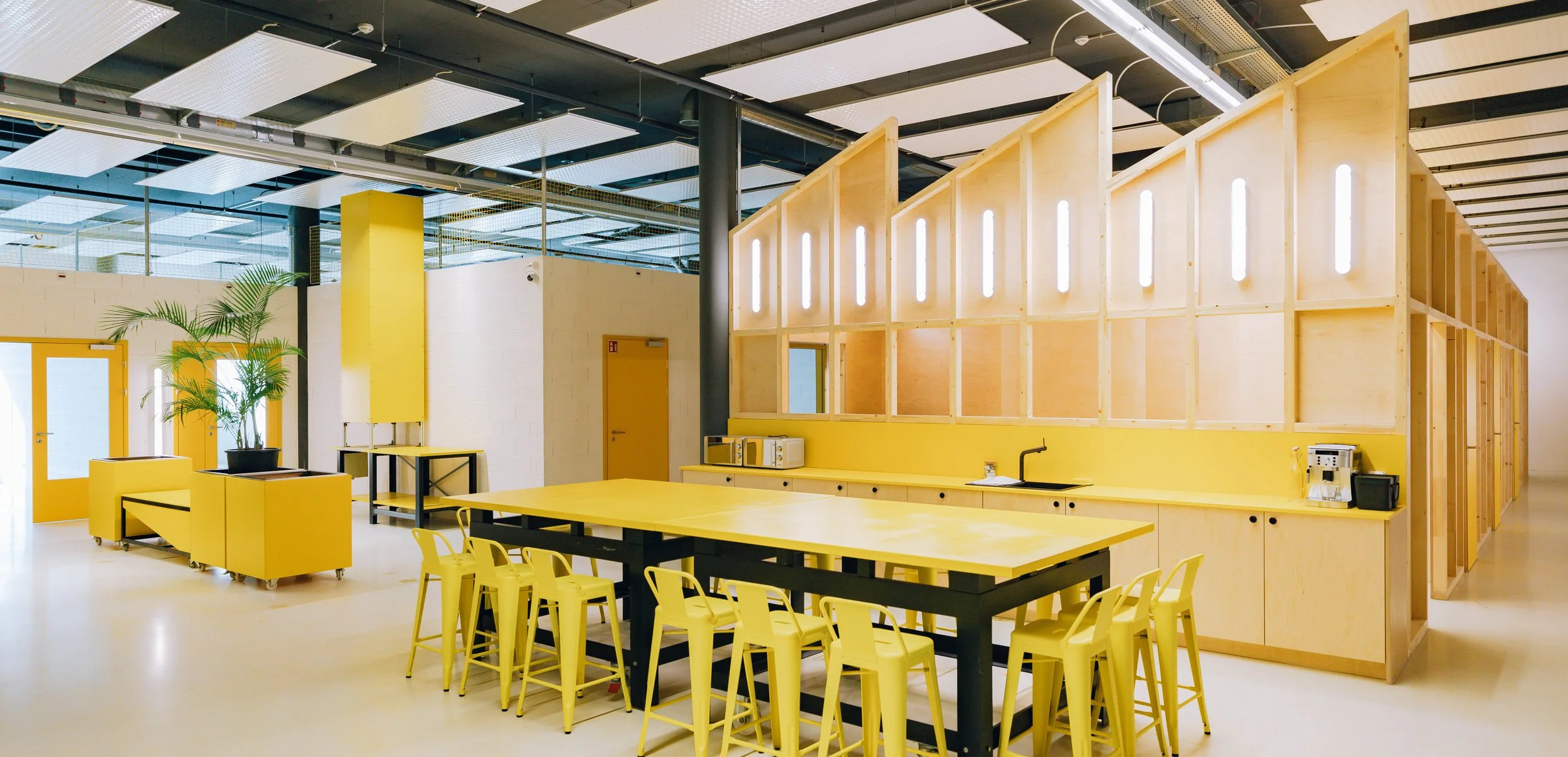 Espace de coworking moderne dans une ancienne usine de panneaux solaires avec cuisine et paroi bois clair, mobilier industriel jaune de réemploi, établis, tabourets, pots de plantes, banc sur roulettes, totem d'accueil, avec lumière naturelle.