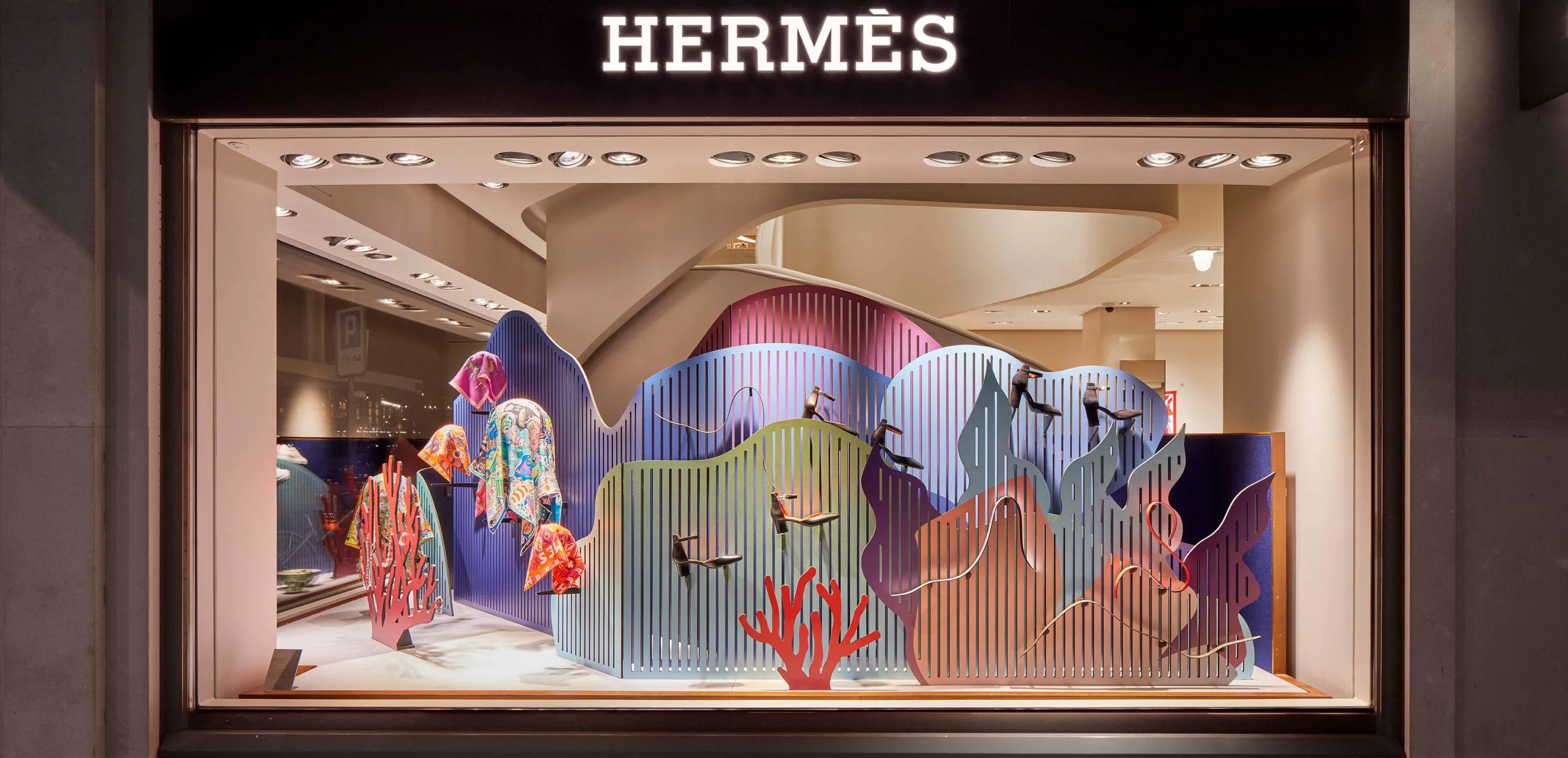 Vitrine d'une boutique Hermès à Genève avec une scénographie colorée de formes ondulées inspirées des fonds marins mettant en valeur des ceintures, chaussures et foulards Hermès, éclairages spot, signature Hermès en haut.