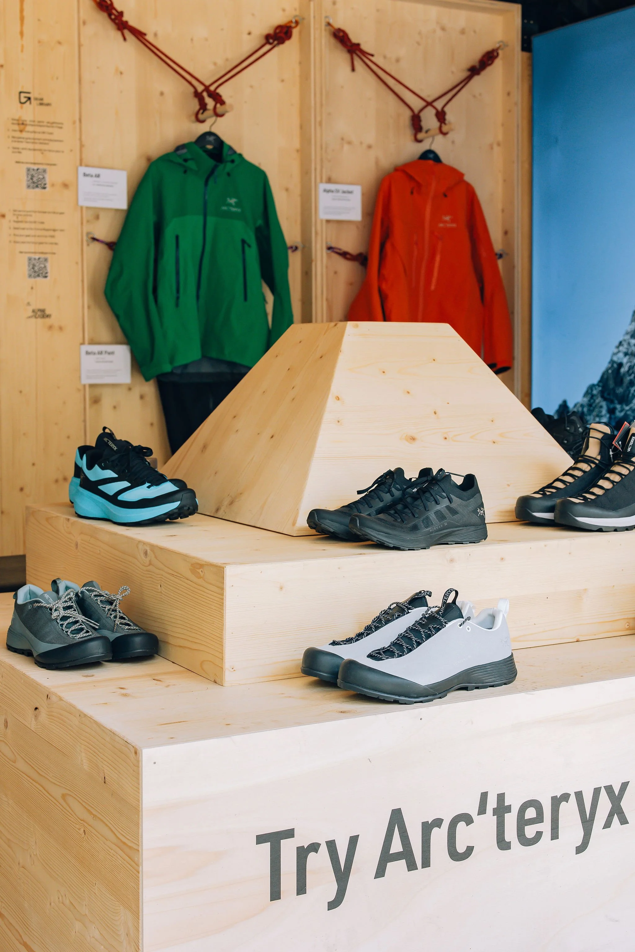 Chaussures d'escalade affichées sur une table en bois dans un magasin d'outdoor, avec des vestes de randonnée vert et orange en arrière-plan.