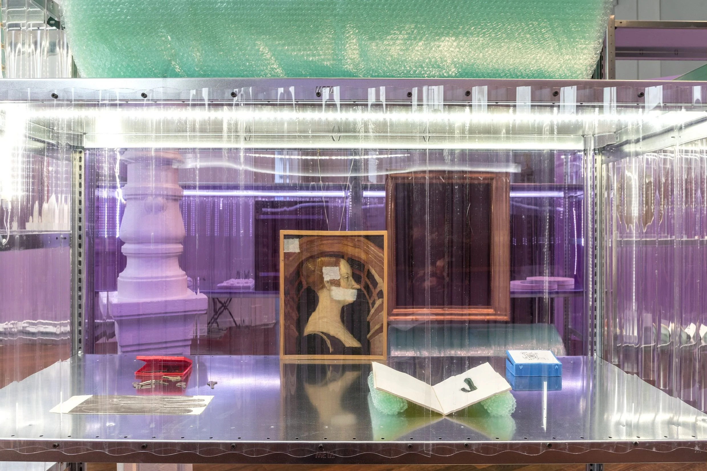 Une vitrine d'exposition contenant divers objets, notamment une peinture, une sculpture en forme de colonne, et des petits accessoires en dessous, avec un éclairage lumineux.