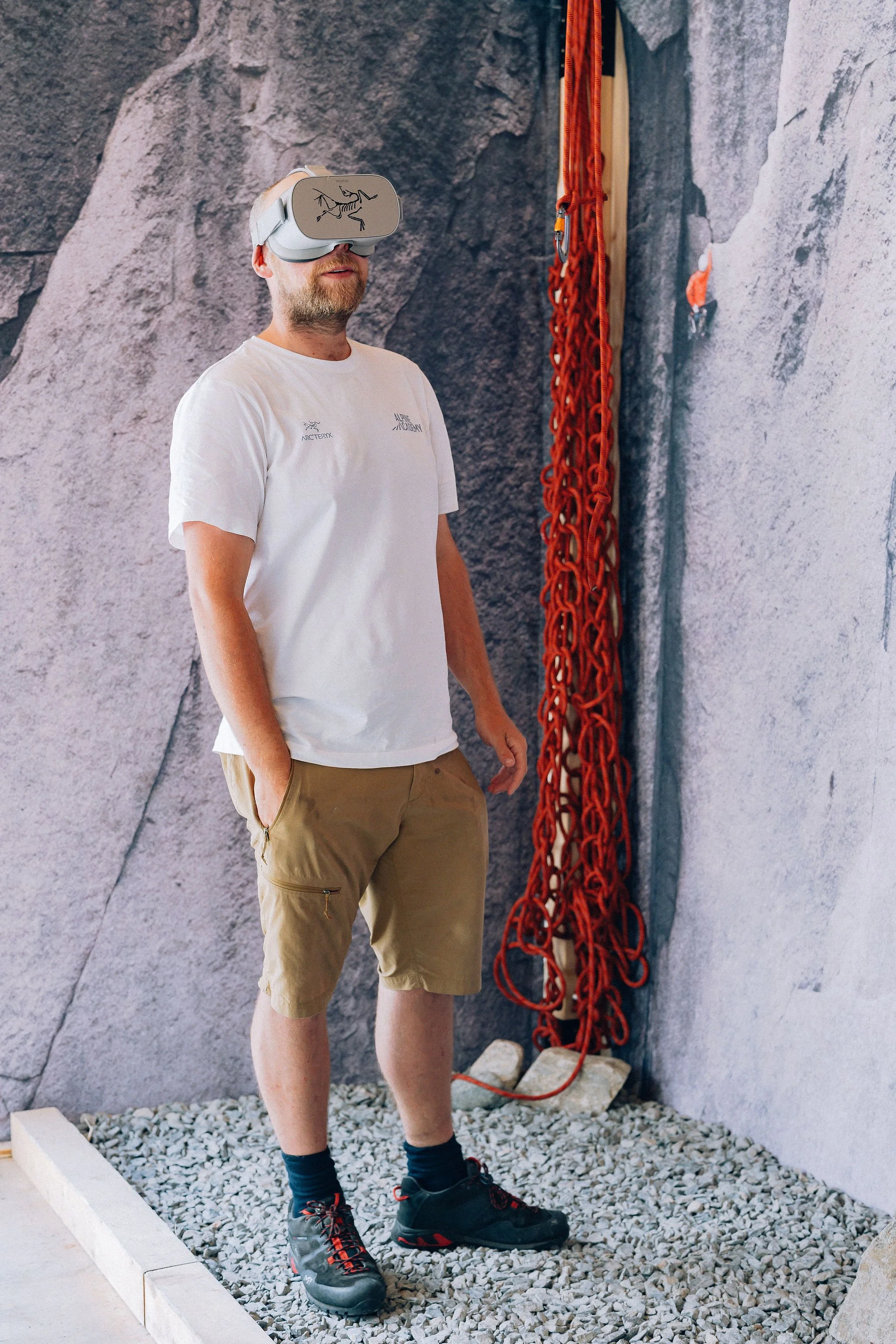 Un homme portant un casque de réalité virtuelle regarde vers le haut dans un espace intérieur avec un mur en pierre et une corde d'escalade. Il porte un t-shirt blanc, un pantalon beige, des chaussettes noires et des chaussures de randonnée. Le sol est recouvert de petites pierres.