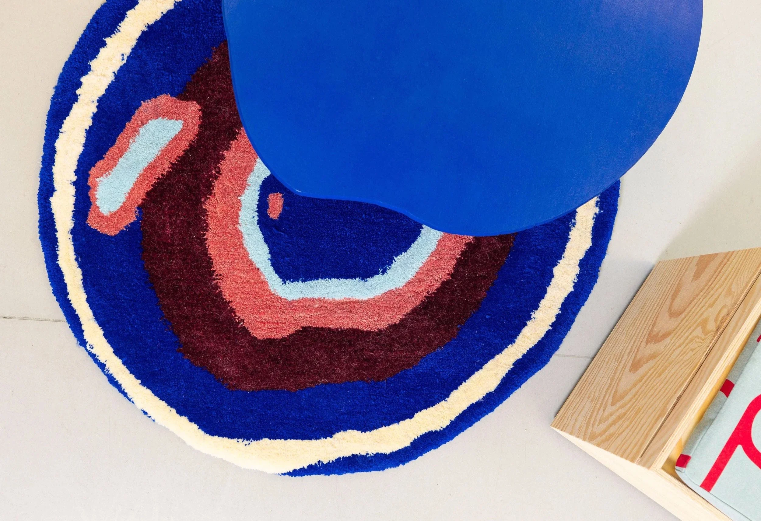 Tapis en forme d'arc-en-ciel avec des couleurs vives et un motif concentrique, posé sur un sol en carrelage blanc, avec deux blocs en bois à droite.