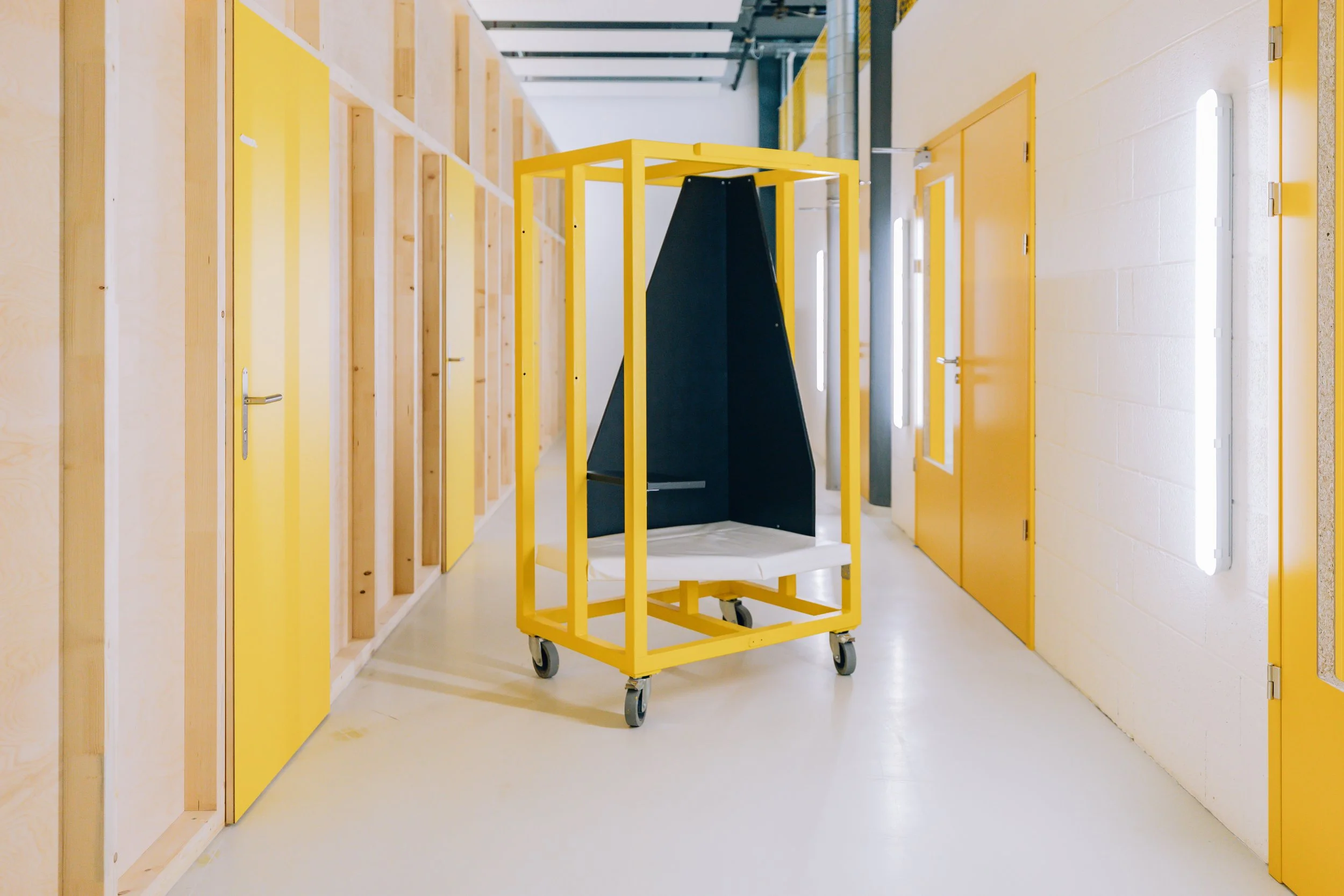 Un chariot de transport jaune avec une plateforme en dessous, situé dans un couloir avec des murs peints en blanc et des portes jaunes, dans un environnement industriel ou d'entrepôt.