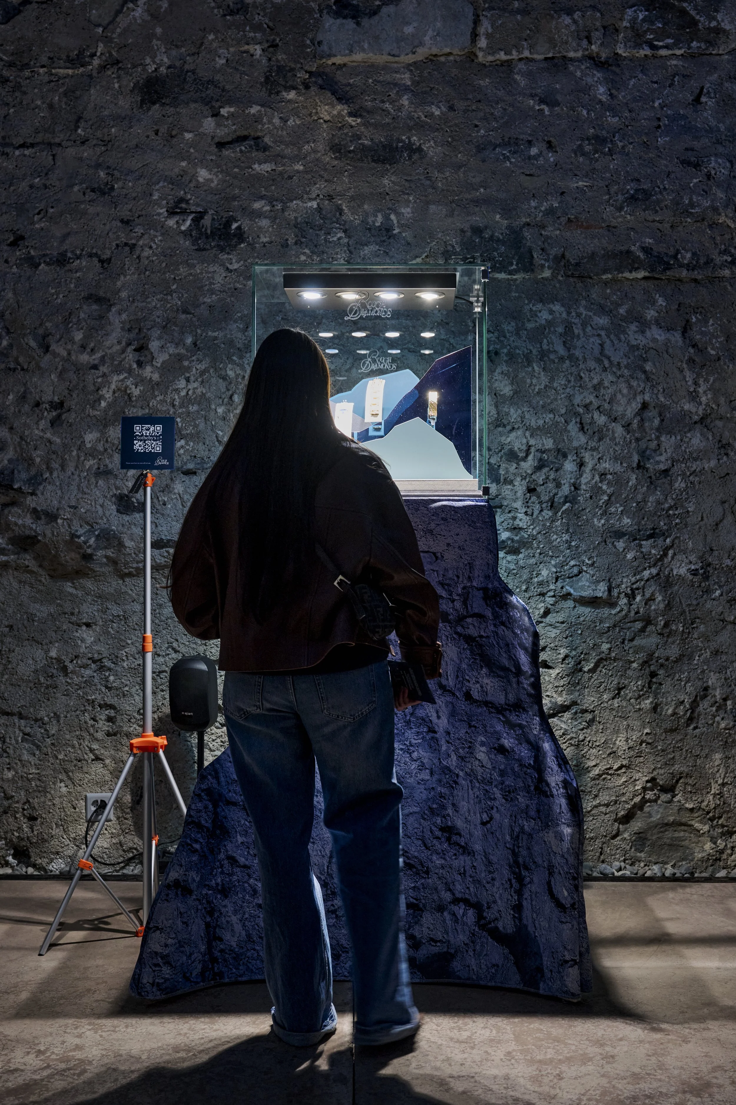 Une femme vue de dos dans une galerie d'exposition, regardant une vitrine contenant des bijoux en diamants, avec un éclairage dirigé sur les pièces, à côté d'une affiche QR code.