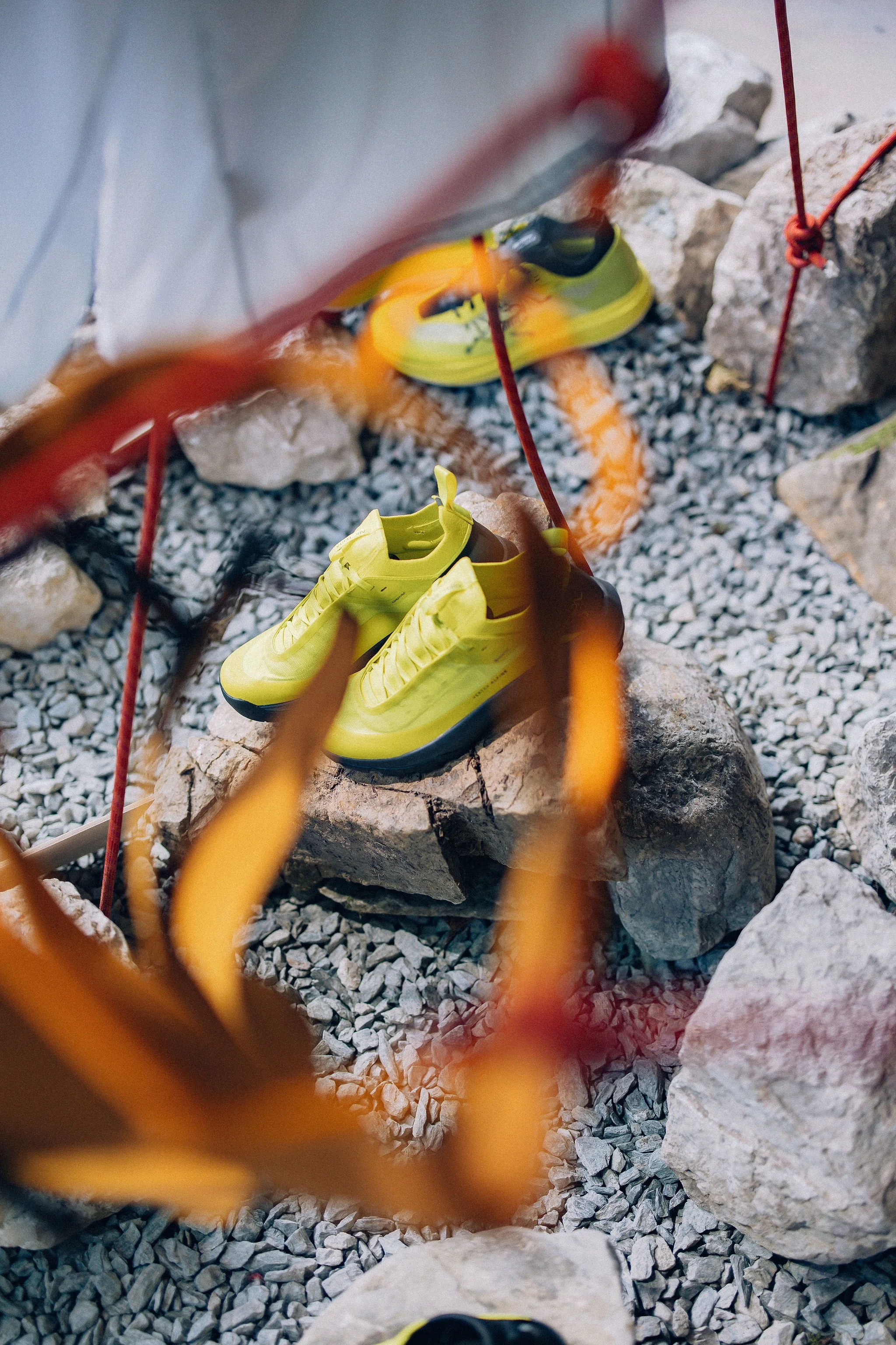 Chaussures de montagne jaune fluo et veste technique blanche de la marque Arc'teryx, mises en valeur dans une scénographie faite de rochers et de graviers, vue en plongée à travers un baudrier orange Arc'teryx.
