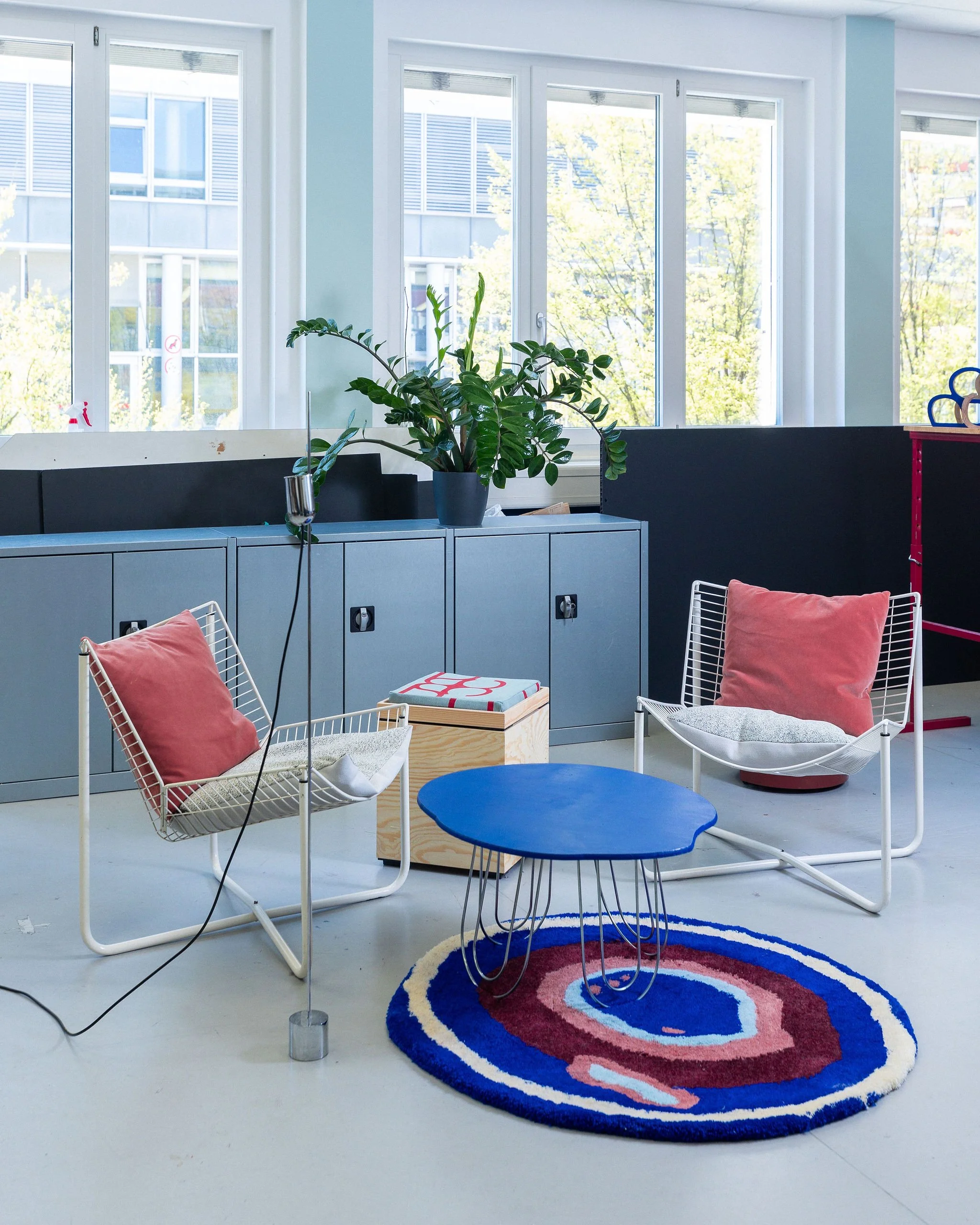 Une salle lumineuse avec de grandes fenêtres, un meuble gris avec une plante verte en pot, plusieurs chaises blanches avec des coussins roses, un tapis rond coloré, et un meuble en bois.