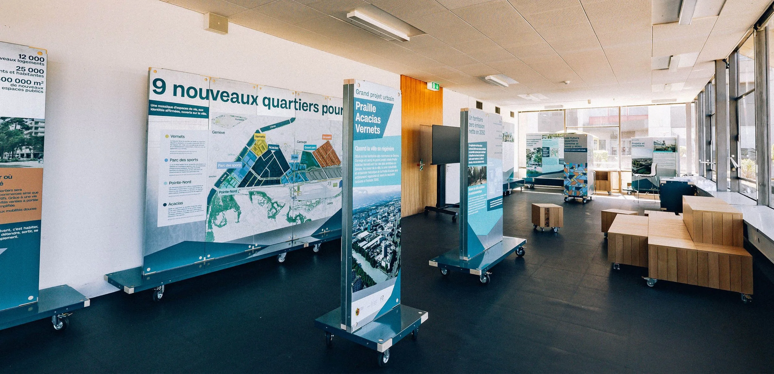 Scénographie d'exposition du projet urbanistique Praille Acacias Vernet à Genève. Salle d'exposition avec panneaux informatifs sur les nouveaux quartiers urbains, des présentoirs mobiles, et de grandes fenêtres laissant entrer la lumière naturelle.