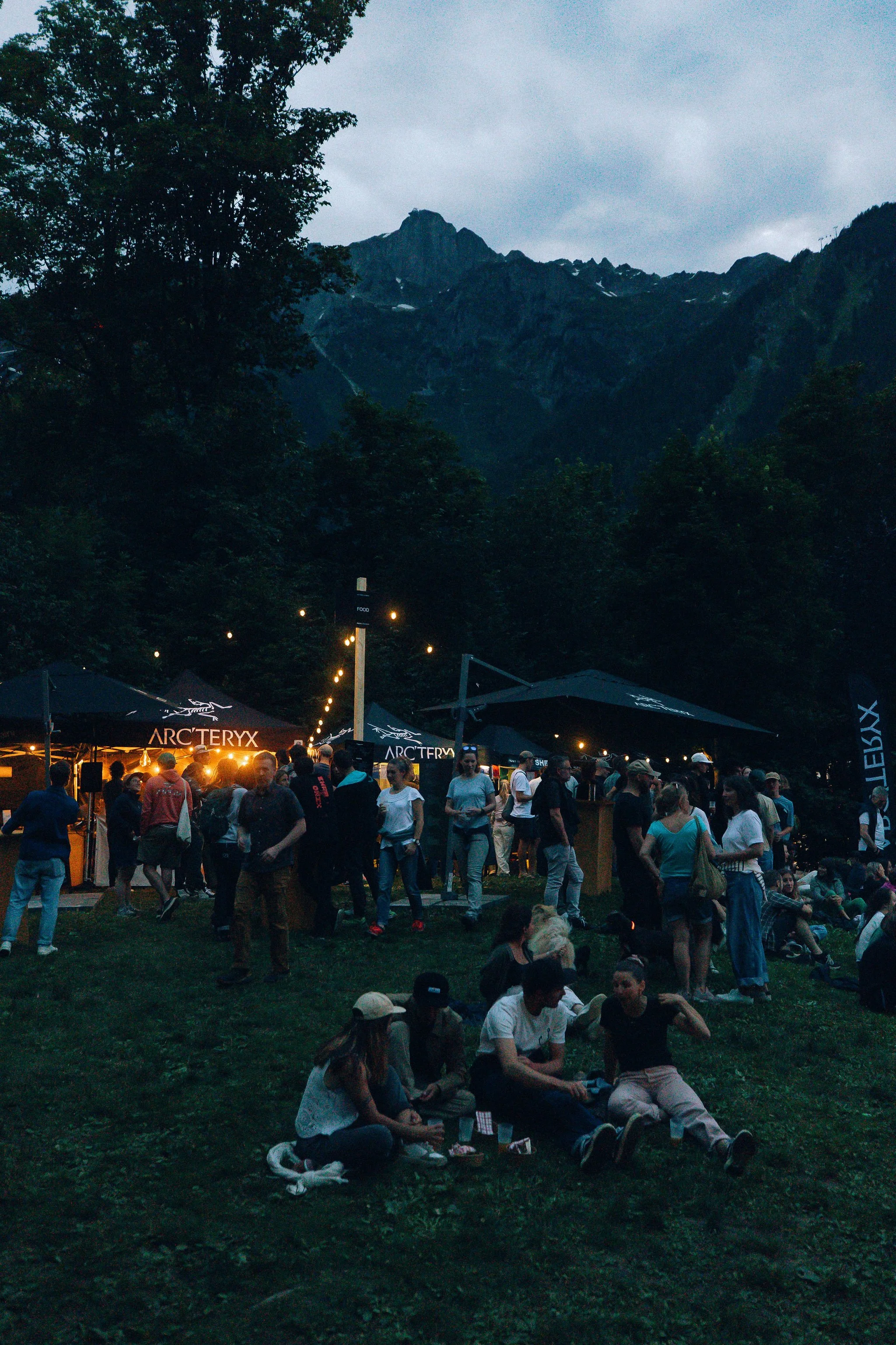 Une foule de personnes se rassemblent dans un festival en plein air la nuit, avec des montagnes en arrière-plan, des tentes avec des logos Arc'teryx, et des lumières suspendues. La scène se déroule dans un environnement naturel entouré de forêt.