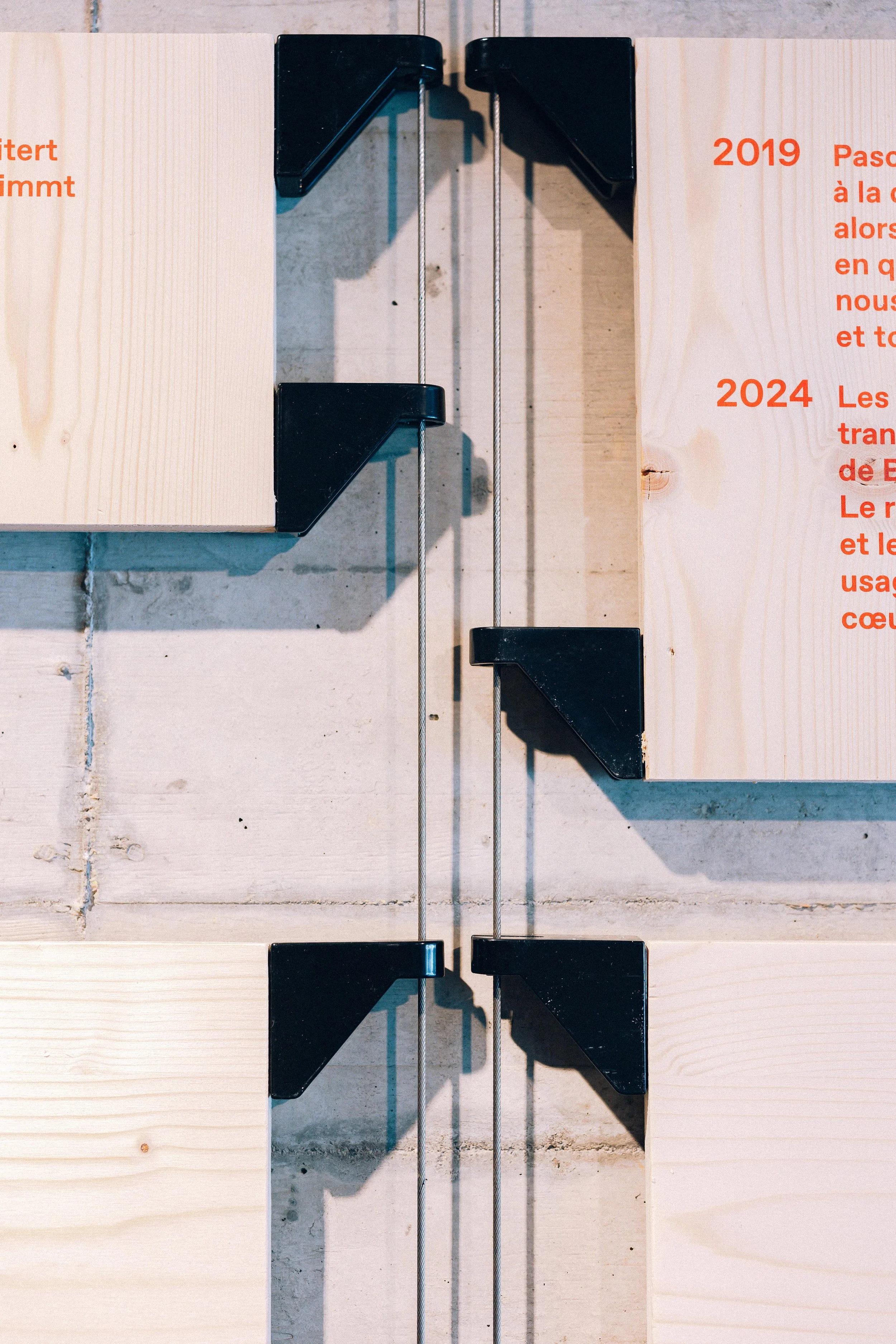 Deux pièces de bois clouées ensemble avec des pinces en plastique noir, suspendues par des câbles métalliques. Le fond est en béton avec une surface en pierre. Il y a du texte en rouge partiellement visible sur le bois.