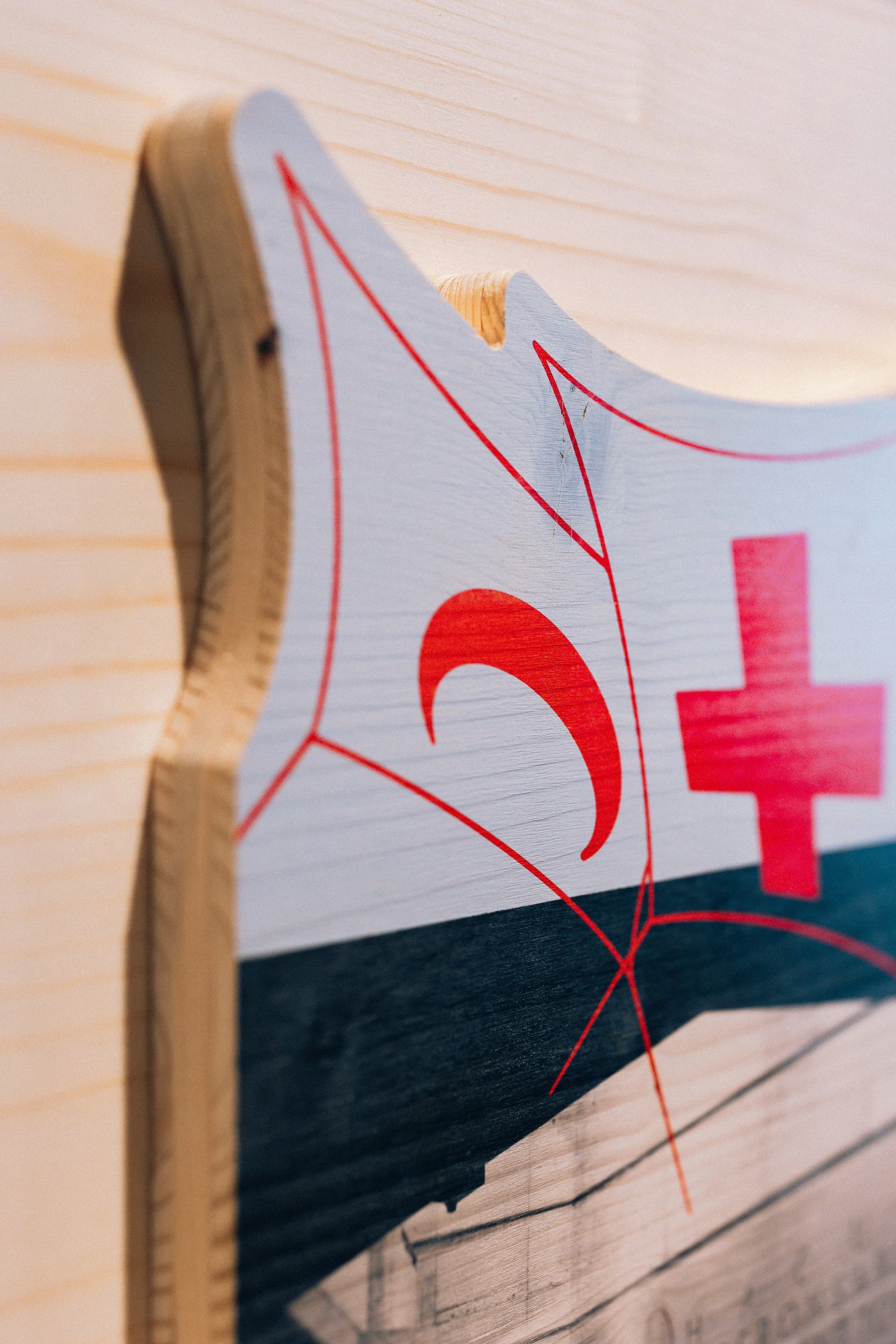 Une planche en bois découpée à la CNC avec une illustration en rouge et blanc représentant le logo du Musée international de la Croix-Rouge, fixée au fond en bois clair de la fresque historique du Musée.