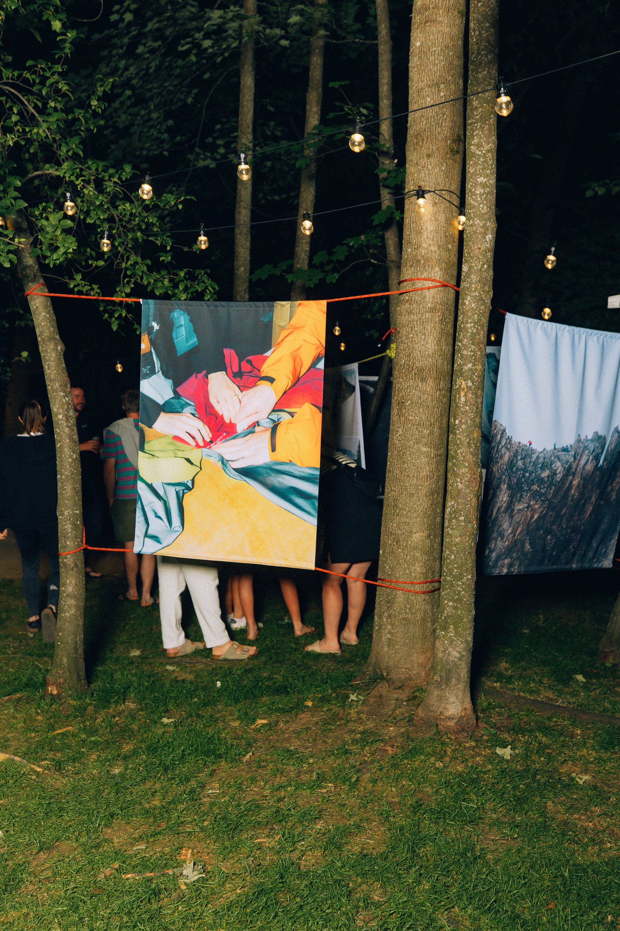 Une scène en plein air dans une forêt avec des arbres et un sol en herbe. Des personnes sont rassemblées en arrière-plan, tandis que plusieurs affiches sont suspendues entre les arbres, représentant des images ou des œuvres d'art. Des guirlandes lumineuses décorent la scène, créant une ambiance festive ou artistique dans la nature.