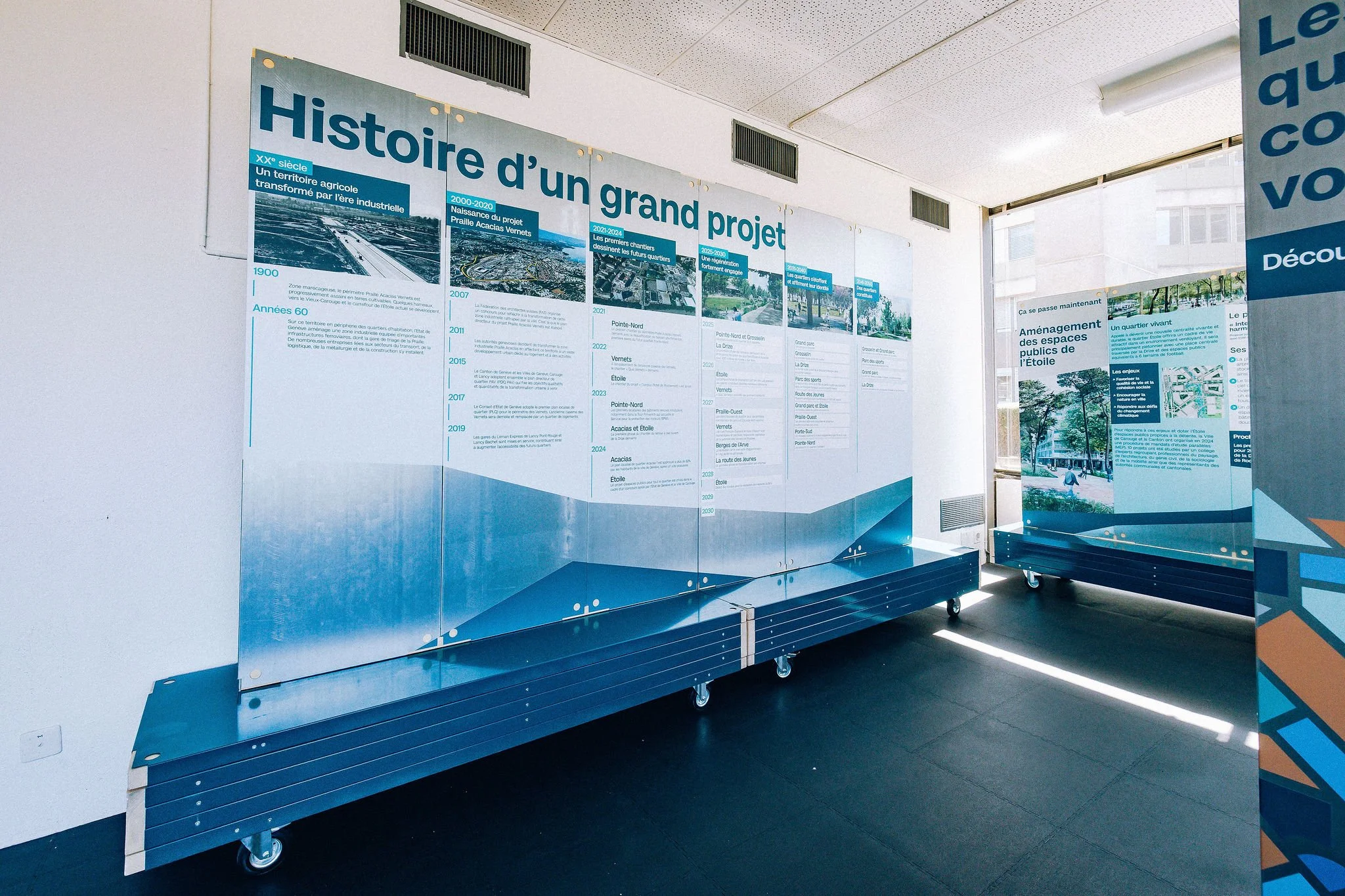 Affichage expliquant l'histoire d'un grand projet urbain, avec plusieurs sections contenant des dates et des descriptions, situé dans une salle lumineuse avec des sièges en arrière-plan.