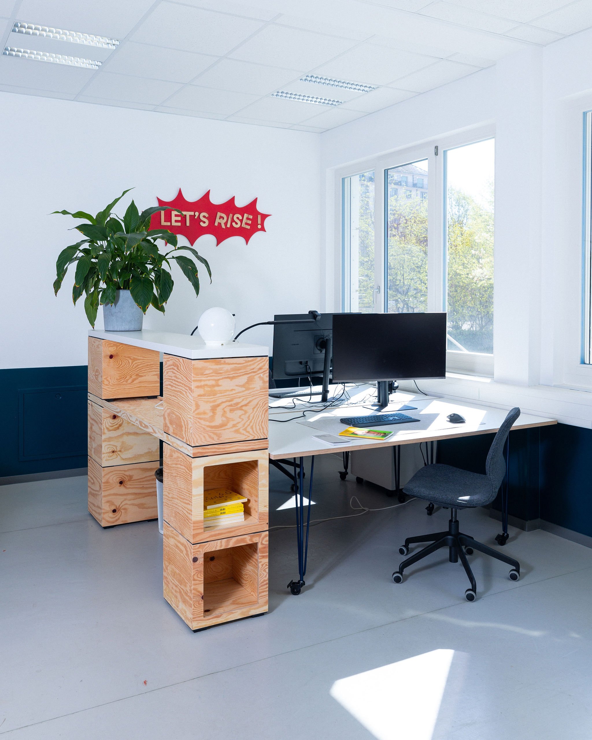 Bureau moderne avec une étagère en bois, une grande plante verte en pot, deux écrans d'ordinateur, une souris, un clavier, et une chaise de bureau. Murs blancs avec une fenêtre laissant entrer de la lumière naturelle. Un panneau rouge avec la phrase "Let's Rise !" est accroché au mur.