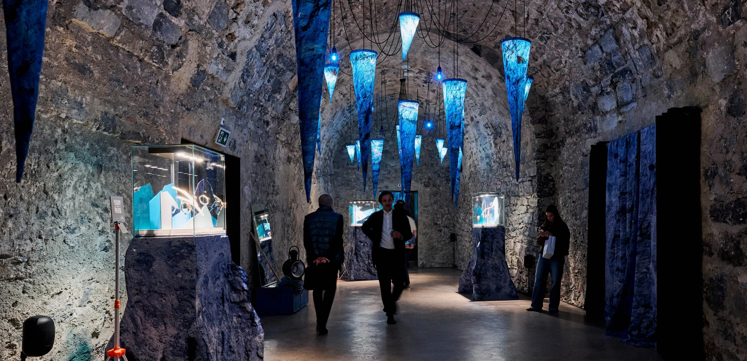 Une exposition dans une cavité rocheuse, avec des pièces d'art ou de design dans des vitrines en verre, illuminée par des lumières bleues et une décoration suspendue en forme de cône. Des visiteurs regardent et discutent au centre, certains utilisant leur téléphone.