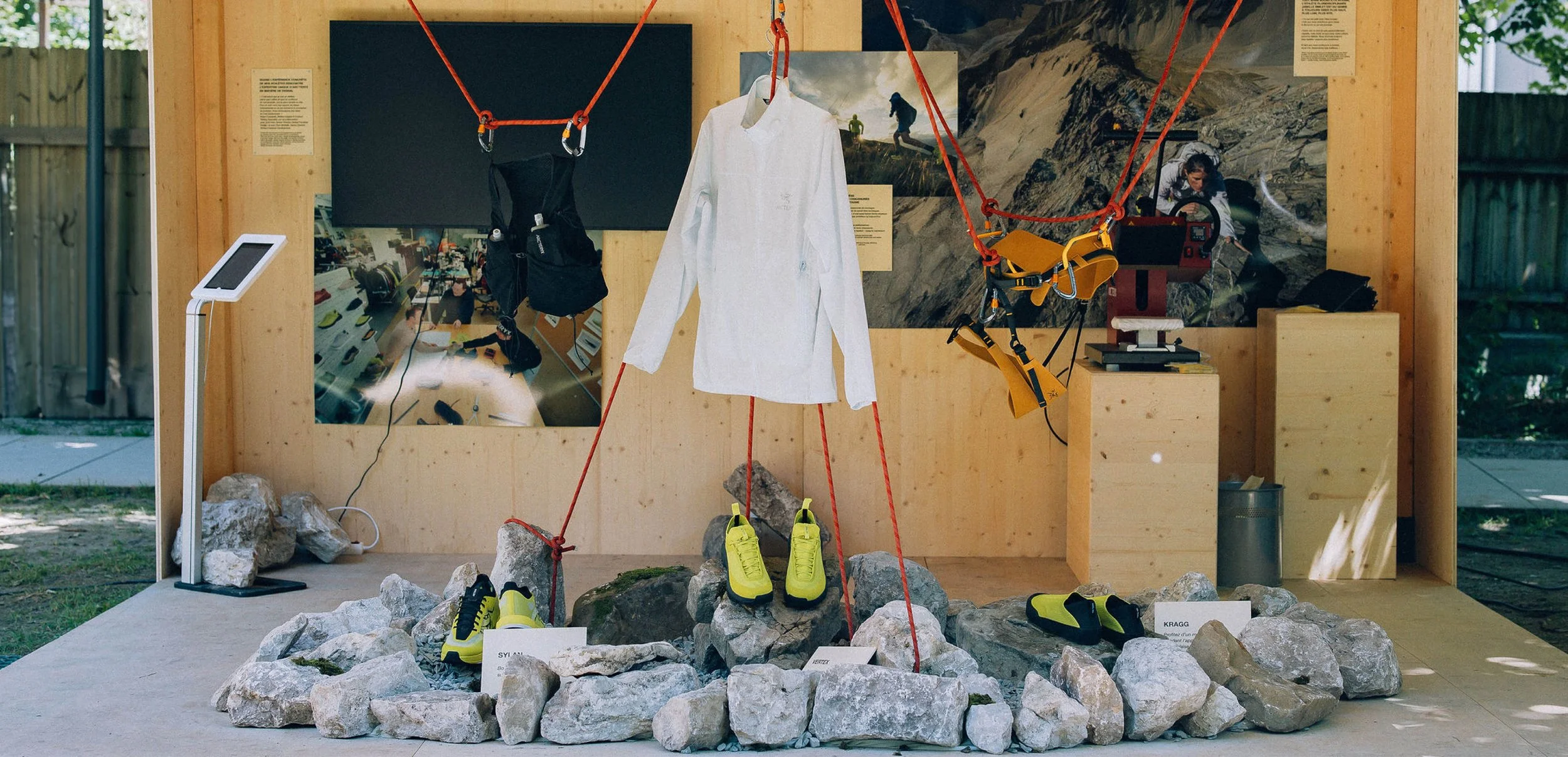 Exposition de vêtements et équipements de montagne, d'alpinisme, d'escalade en plein air, avec chaussures, vêtements techniques, baudrier de la marque Arc'teryx, dans une scénographie composée de rochers, cordes d'escalades et displays en bois.