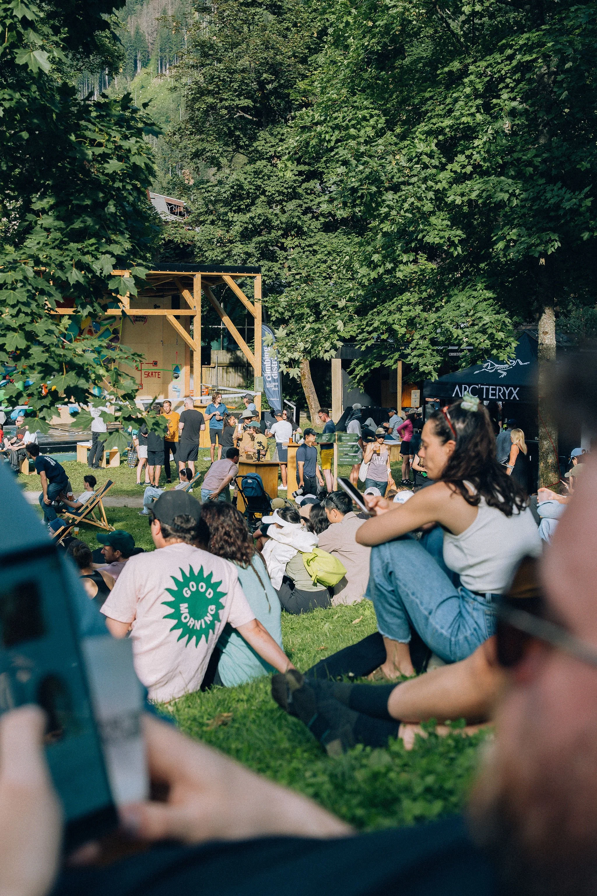 Une foule de personnes assises et debout dans un espace vert entouré d'arbres, lors d'un événement en extérieur. On aperçoit un bâtiment en bois et un stand avec la marque Arc'teryx.