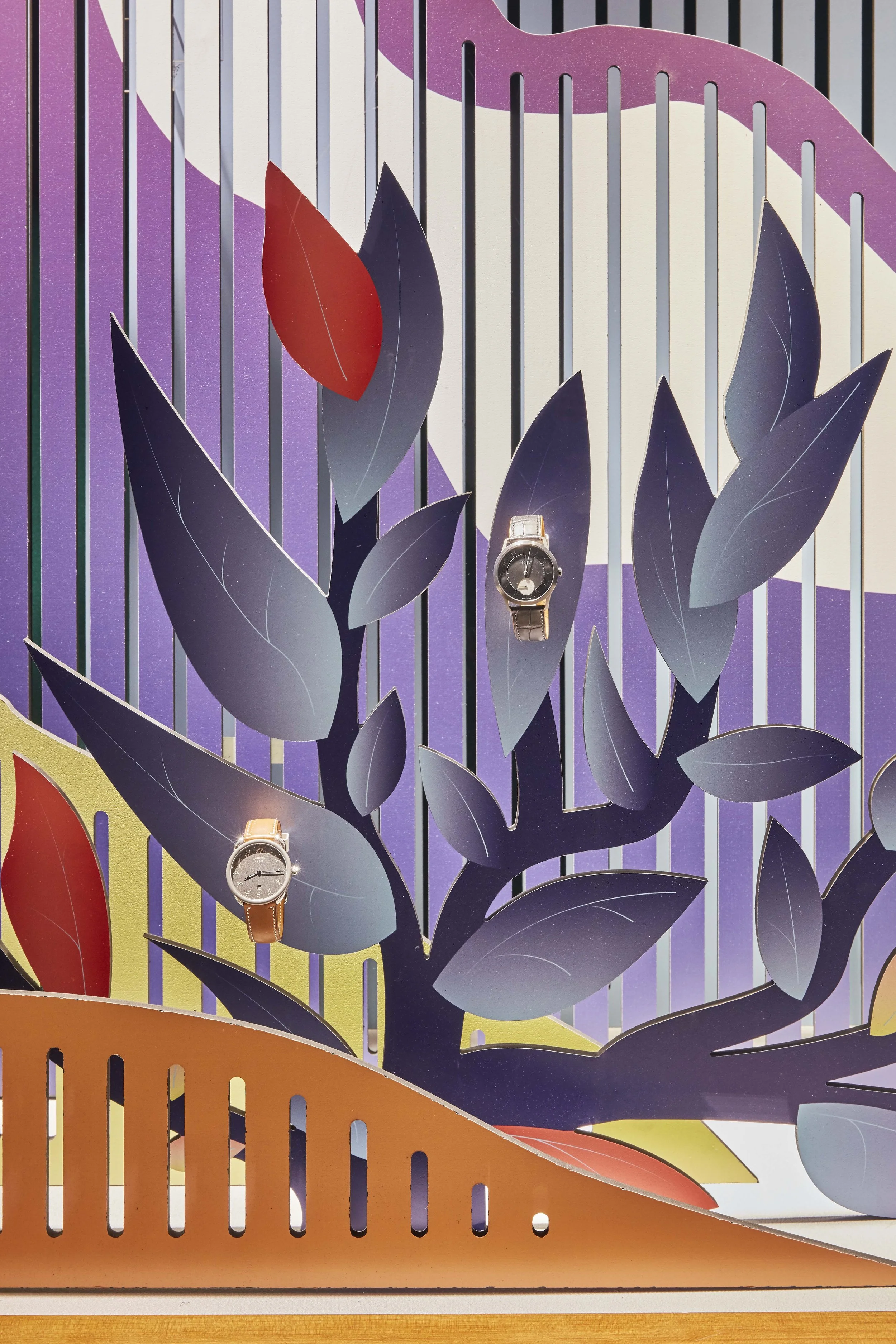 Une vitrine Hermès avec une scénographie composée de feuillages stylisés en couleurs violet, rouge, gris et rose, avec deux montres exposées au centre.