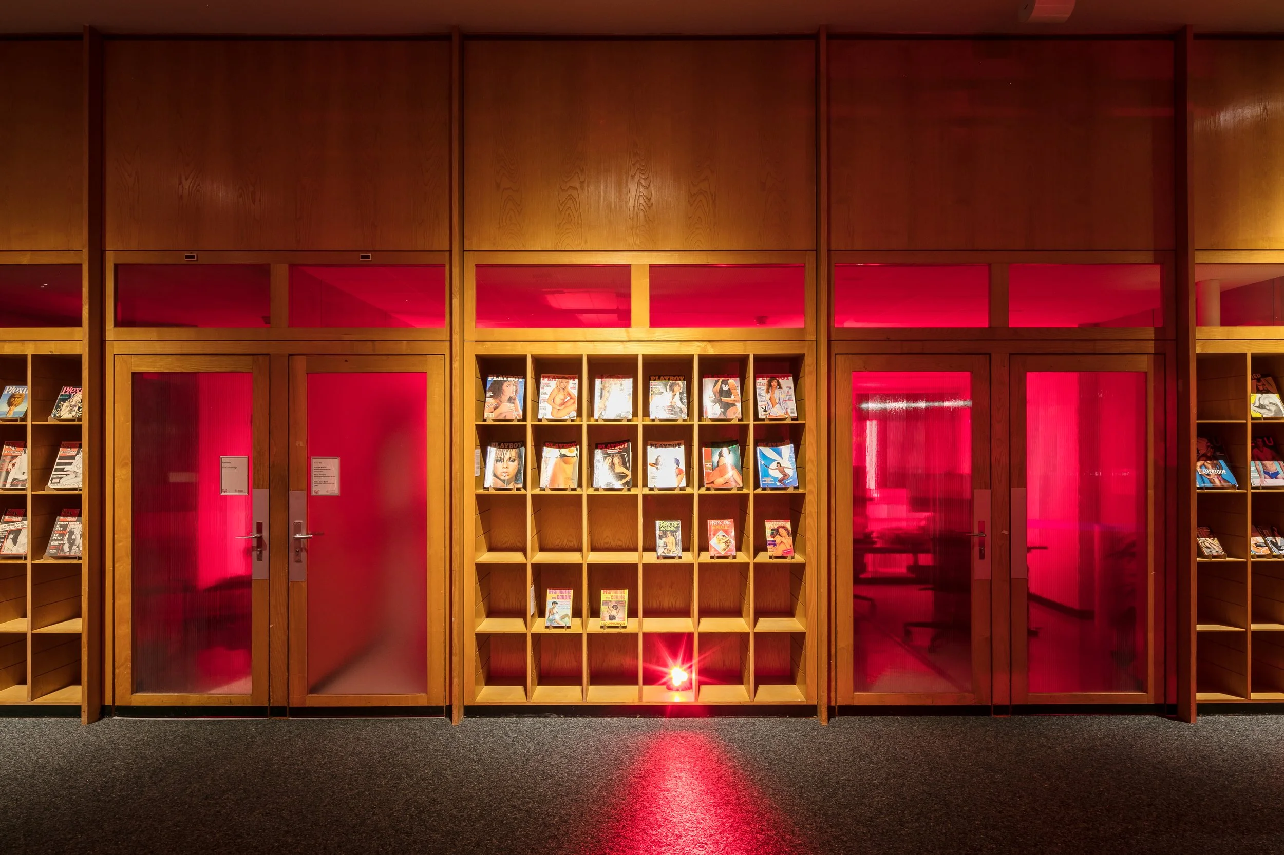 Scénographie d'exposition dans une bibliothèque avec des étagères en bois contenant des magazines Playboy, des lumières rouges rasante, derrière des portes en verre avec lumière rouge à l'intérieur et un mur fait de panneaux de bois.