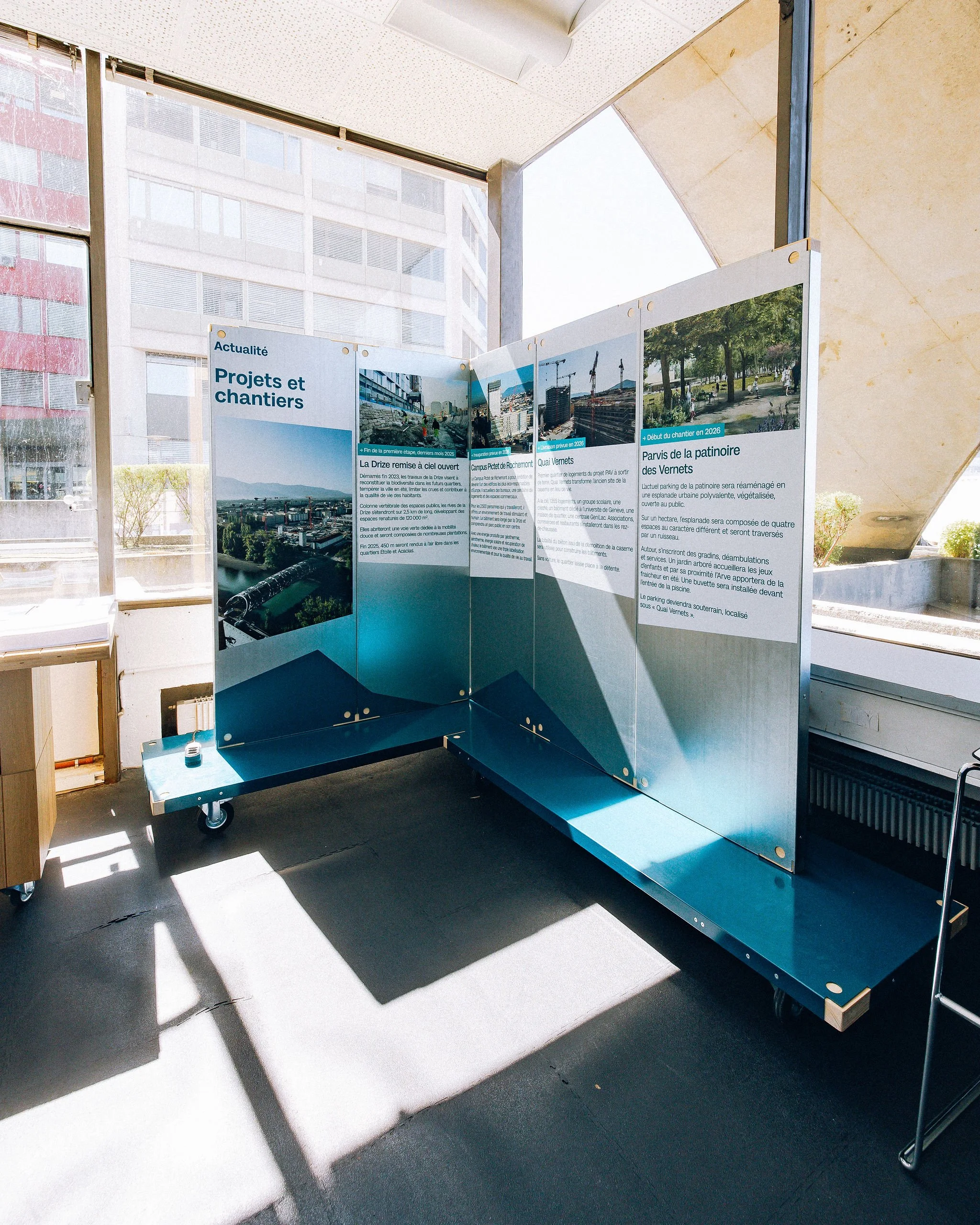 Scénographie d'exposition du projet urbanistique Praille Acacias Vernet à Genève. Une affiche d'informations sur des projets et chantiers de construction, placée dans un espace intérieur avec de grandes fenêtres laissant passer la lumière du jour.
