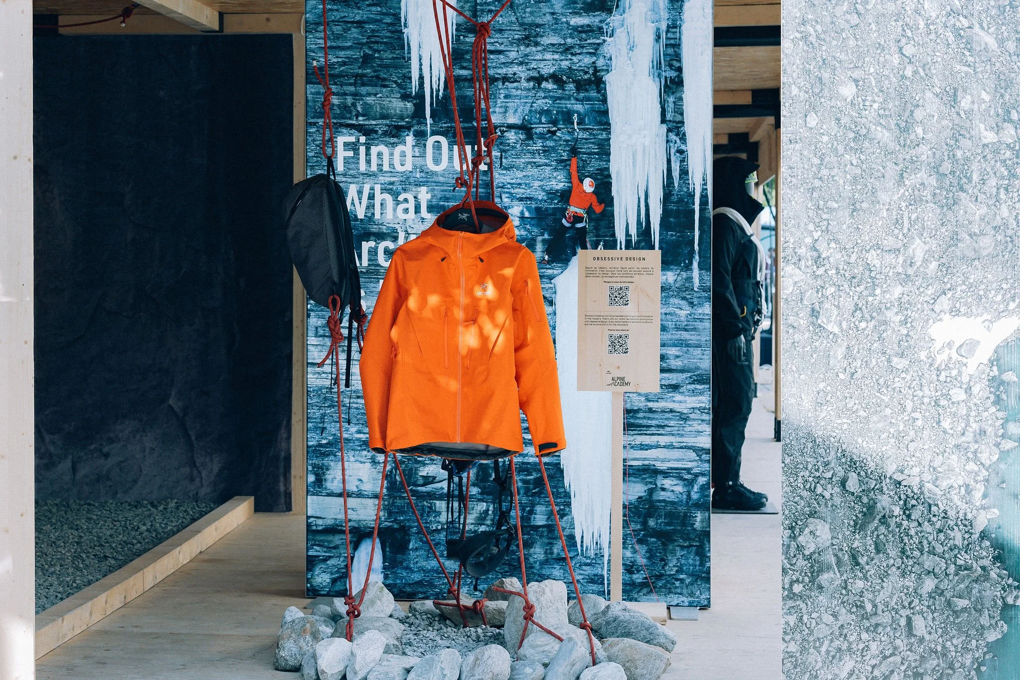 Vitrine d'une boutique d'escalade avec un manteau orange, une affiche sur la gauche mentionnant 'Find Out What', une figurine d'alpiniste, des cordes rouges, une colline avec des pierres au sol, et une personne portant un sac à dos dans le fond.