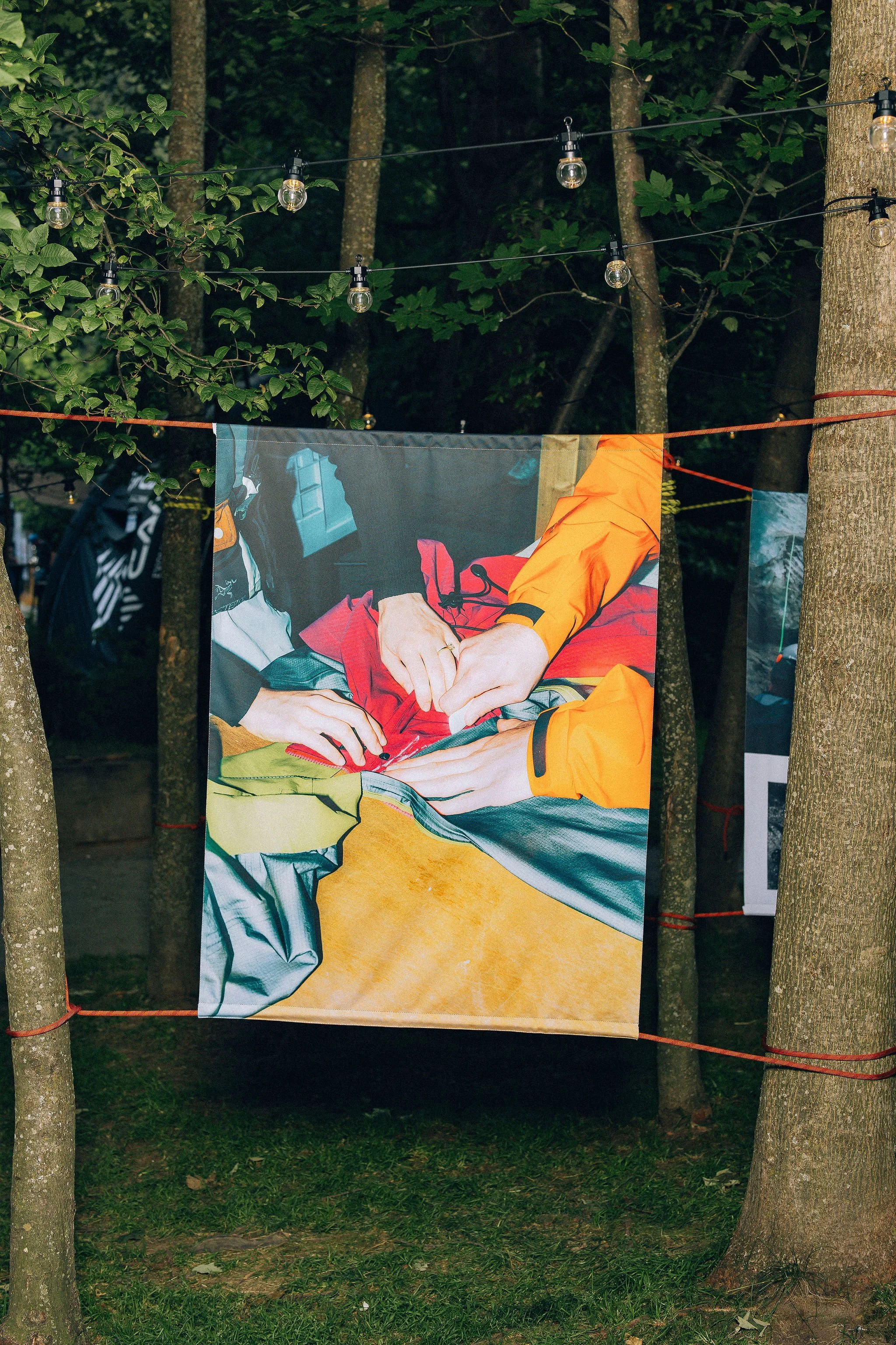 Exposition de photos de la marque Arc'teryx pendant le festival Alpine Academy, photo suspendue par des cordes d'escalade entre deux arbres dans un parc, représentant des personnes fabriquant des vêtements orange et gris.