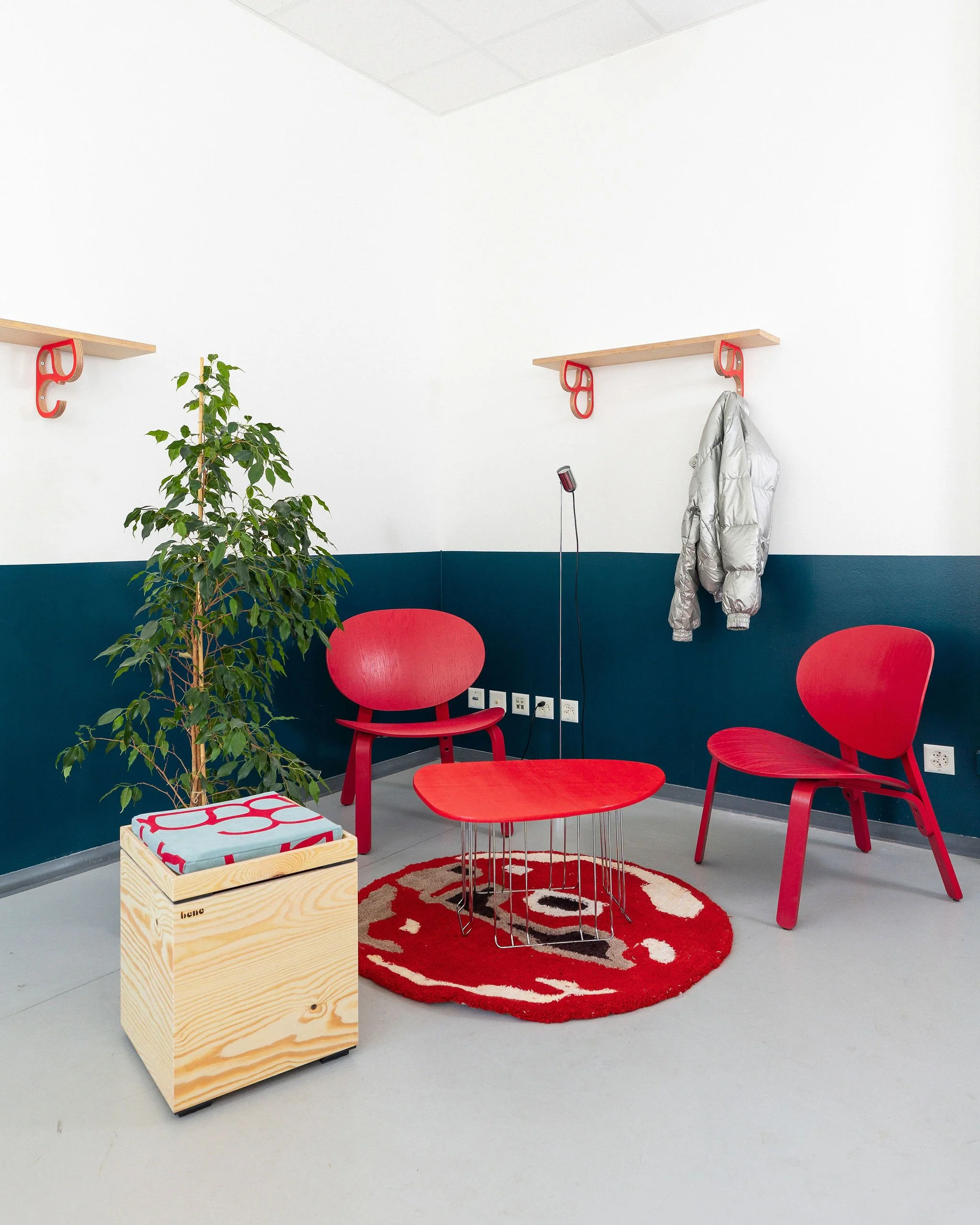 Une salle d'attente moderne avec deux chaises rouges, une table en fil métallique en forme de cylindre avec une bibliothèque en bois en forme de cube, un tapis rond rouge avec un motif blanc, un mur blanc avec une partie basse colorée en bleu foncé, une plante verte, un manteau gris suspendu sur un crochet, une lampe de bureau noire, des prises électriques et une multiprise, deux étagères en bois fixées au mur avec des crochets rouges.