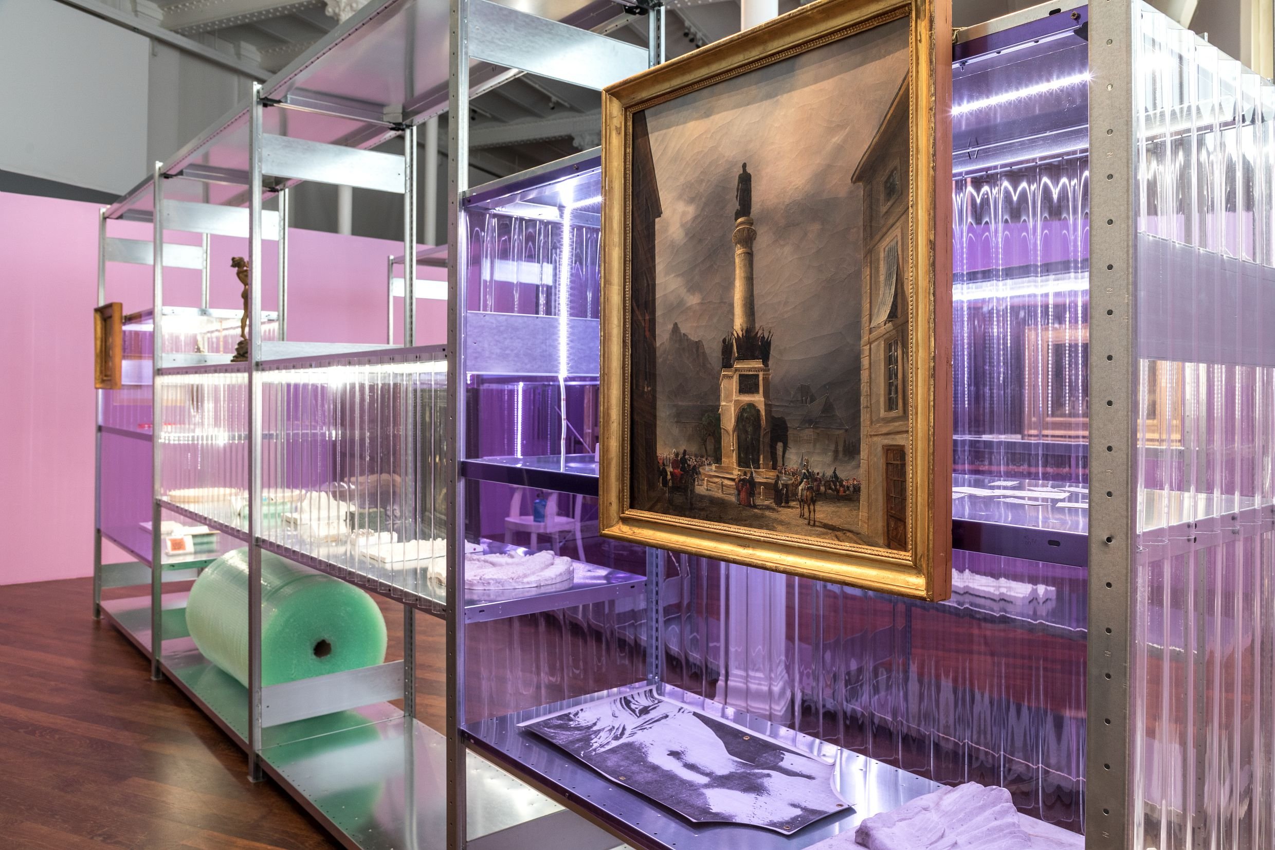 Une structure en métal avec des compartiments en plastique violet contenant des objets variés, dont une peinture encadrée représentant une monument et plusieurs ballons de différentes couleurs