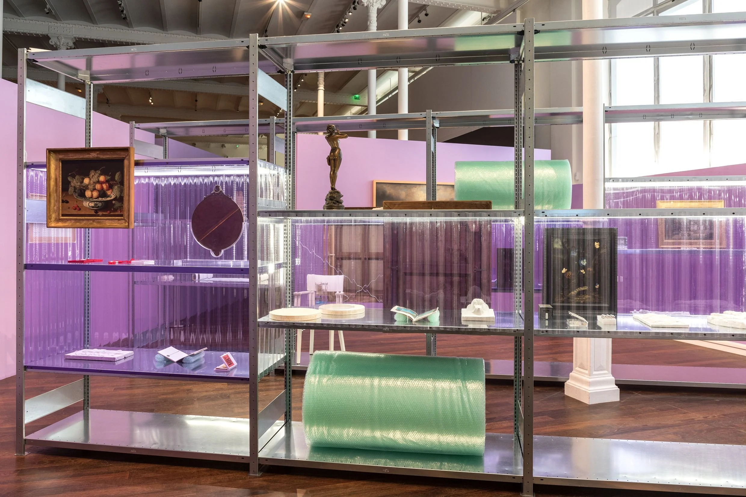 Une étagère d'exposition en métal avec plusieurs niveaux, contenant diverses œuvres d'art et objets, dans une salle d'exposition lumineuse avec des murs violets et des fenêtres