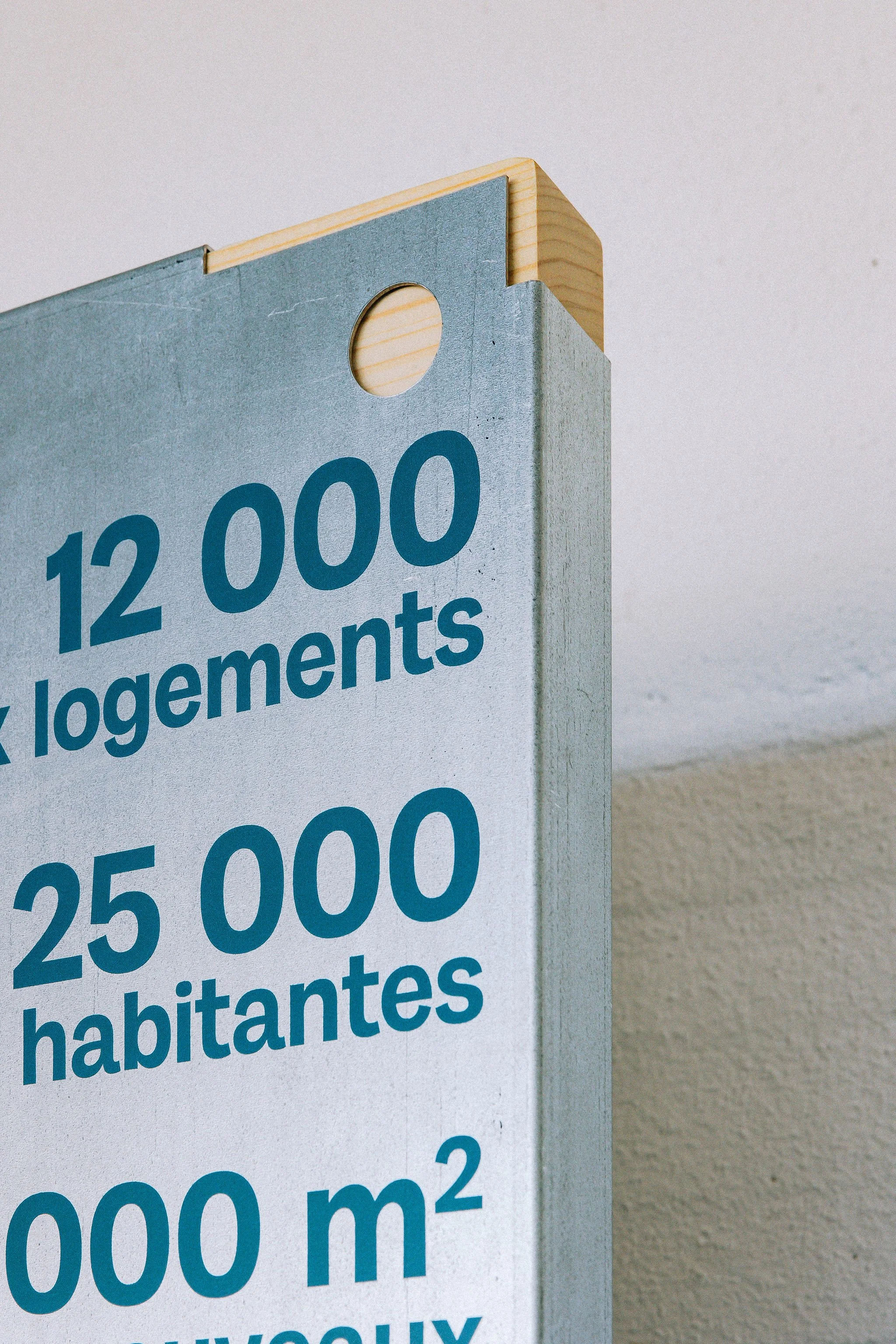 Scénographie d'exposition du projet urbanistique Praille Acacias Vernet à Genève. Panneau d'information en métal brut sur fond de bois avec des chiffres, mentionnant 12 000 logements, 25 000 habitants, et 2000 m²,