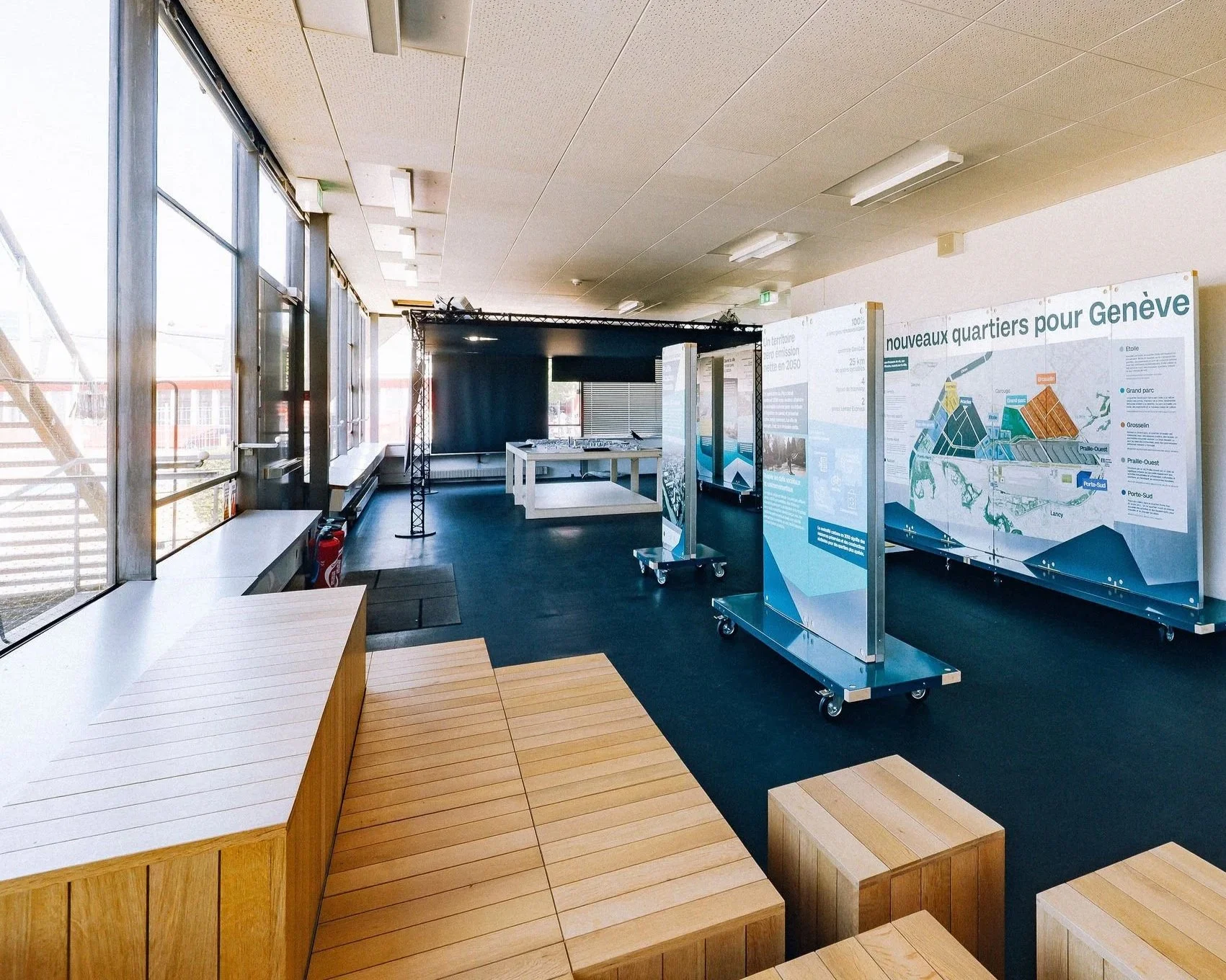 Salle d'exposition avec panneaux d'information sur des quartiers de Genève, grande fenêtre laissant entrer la lumière naturelle, espace avec mobilier en bois, structures temporaires pour présentation.