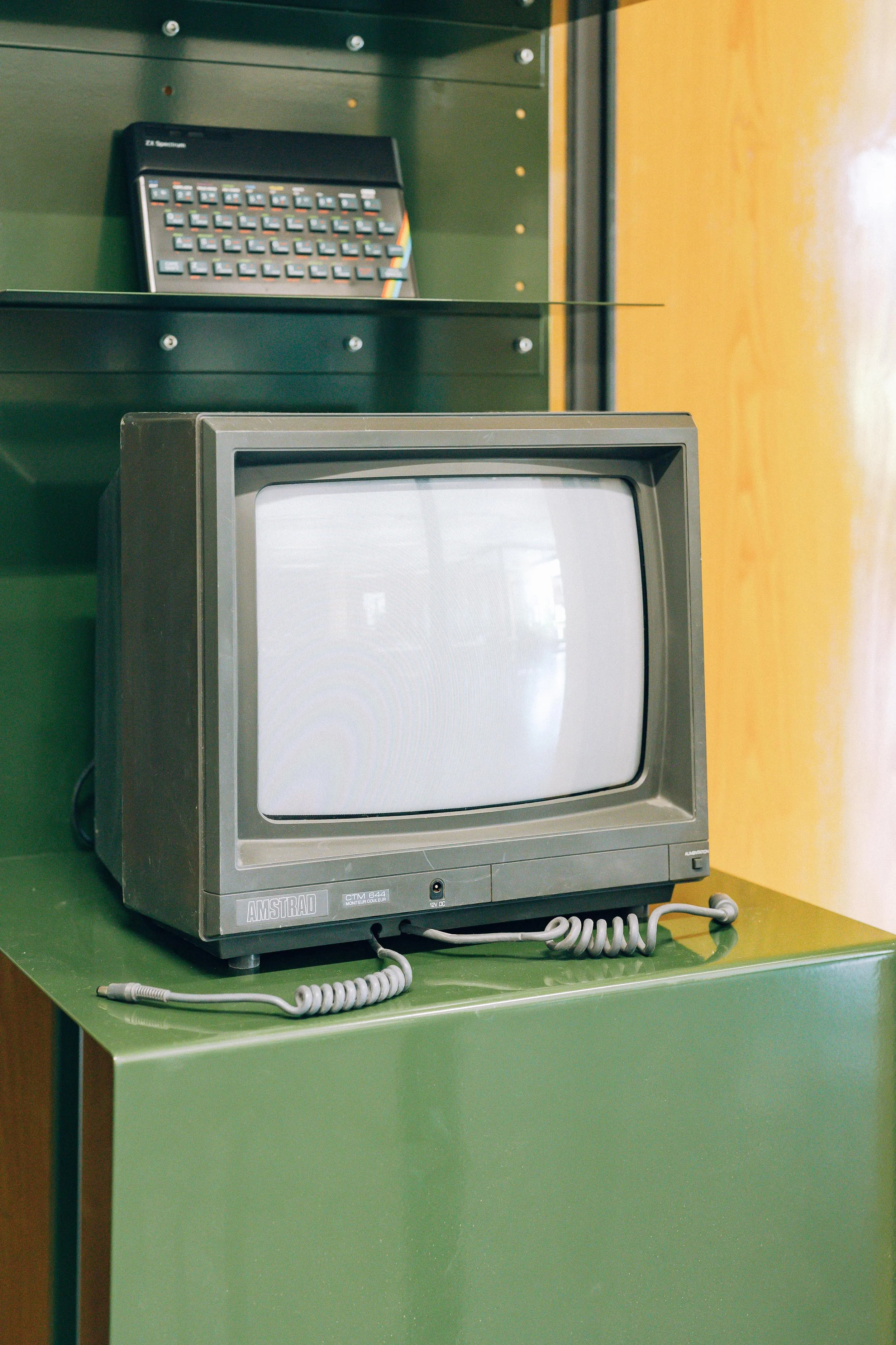 Une télévision rétro en couleur gris argenté posée sur un meuble vert, avec un téléphone à cordon gris dessus, dans un décor intérieur avec un mur vert et une surface en bois au fond.