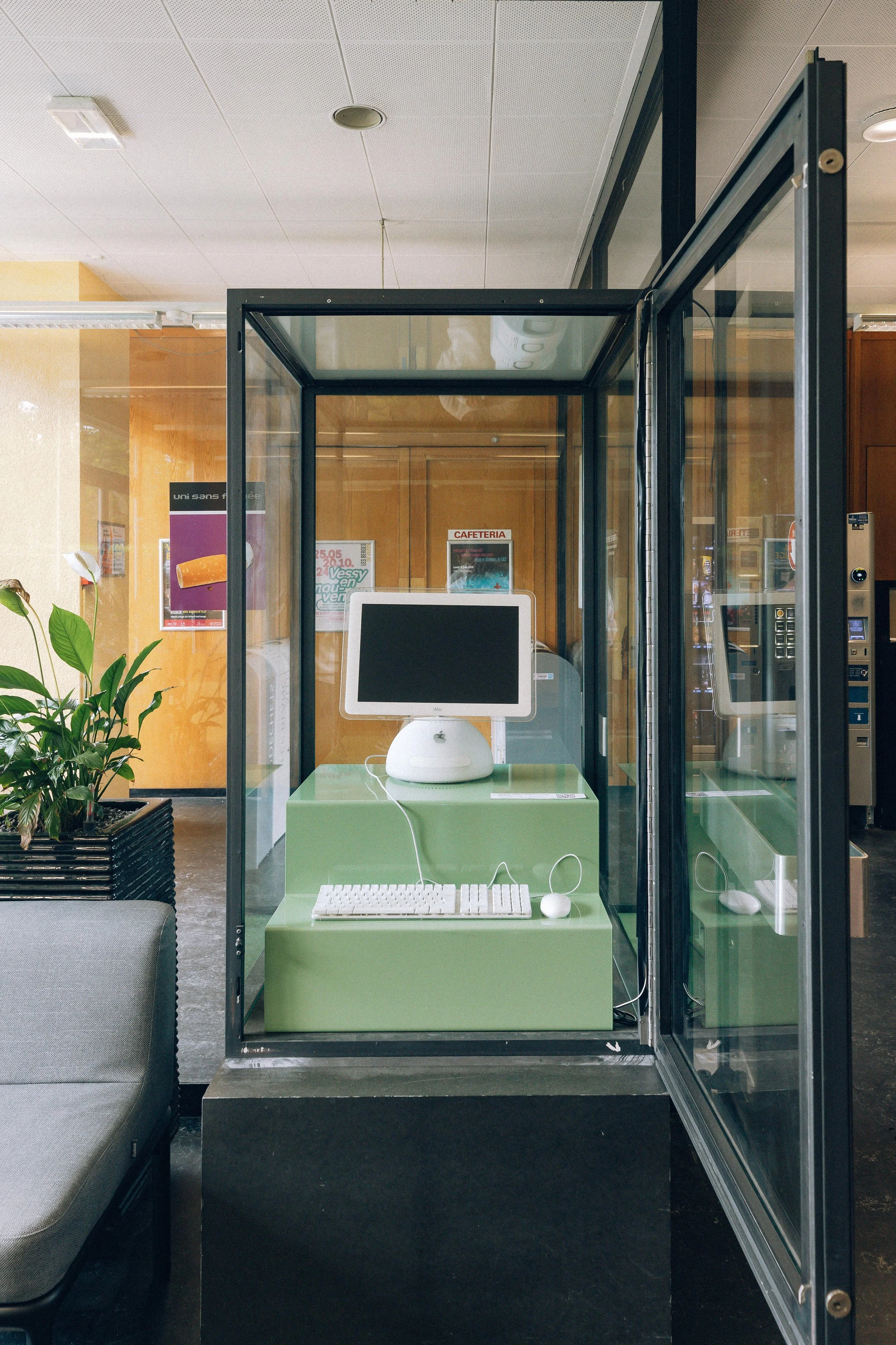 Un poste informatique avec un écran, un clavier, une souris et un casque dans une cabine en verre dans un espace intérieur moderne.