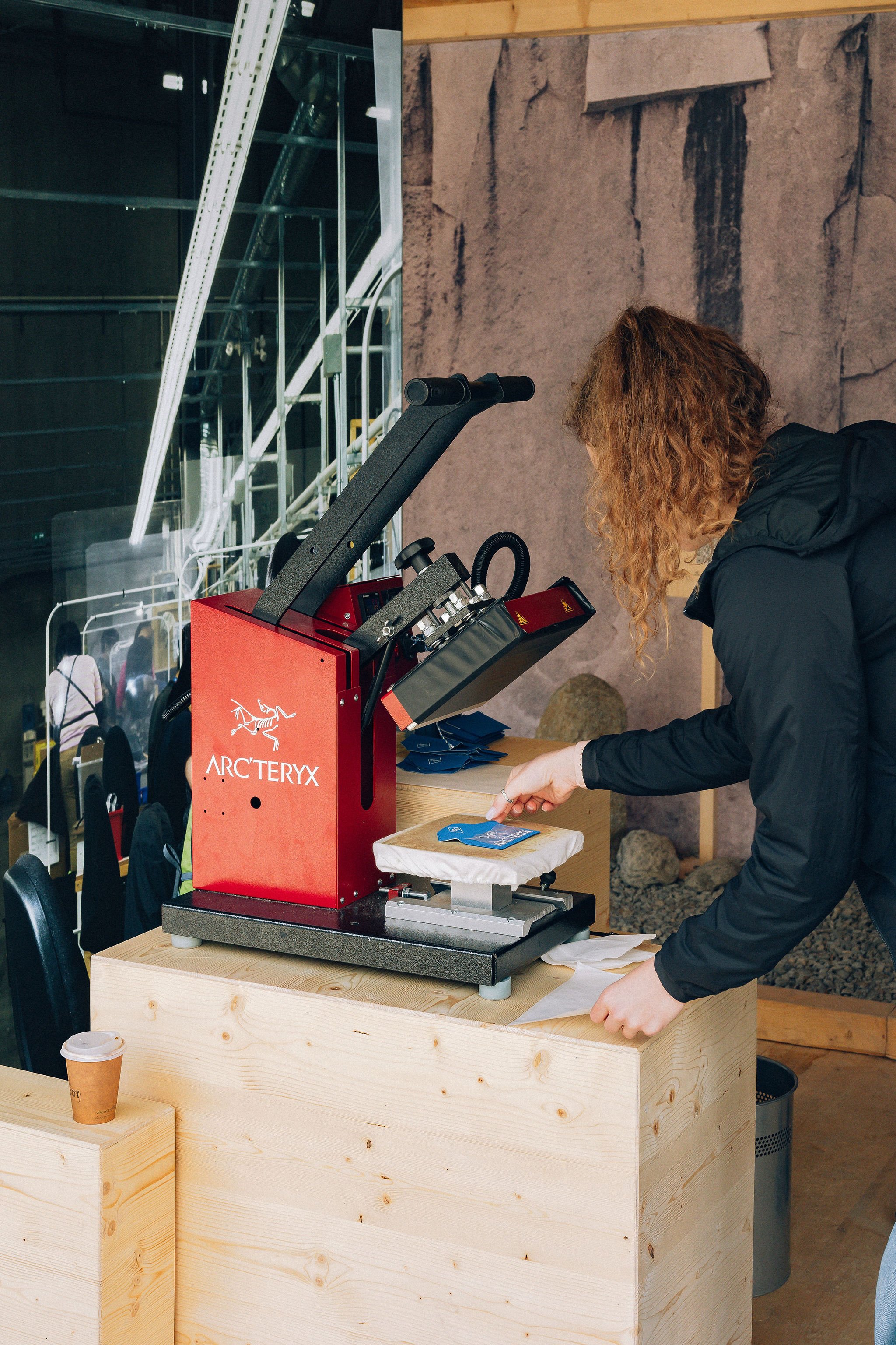 Une personne portant une veste noire regarde une machine à tickets orange avec le logo ARC'TERYX, sur une table en bois claire, dans un environnement intérieur.