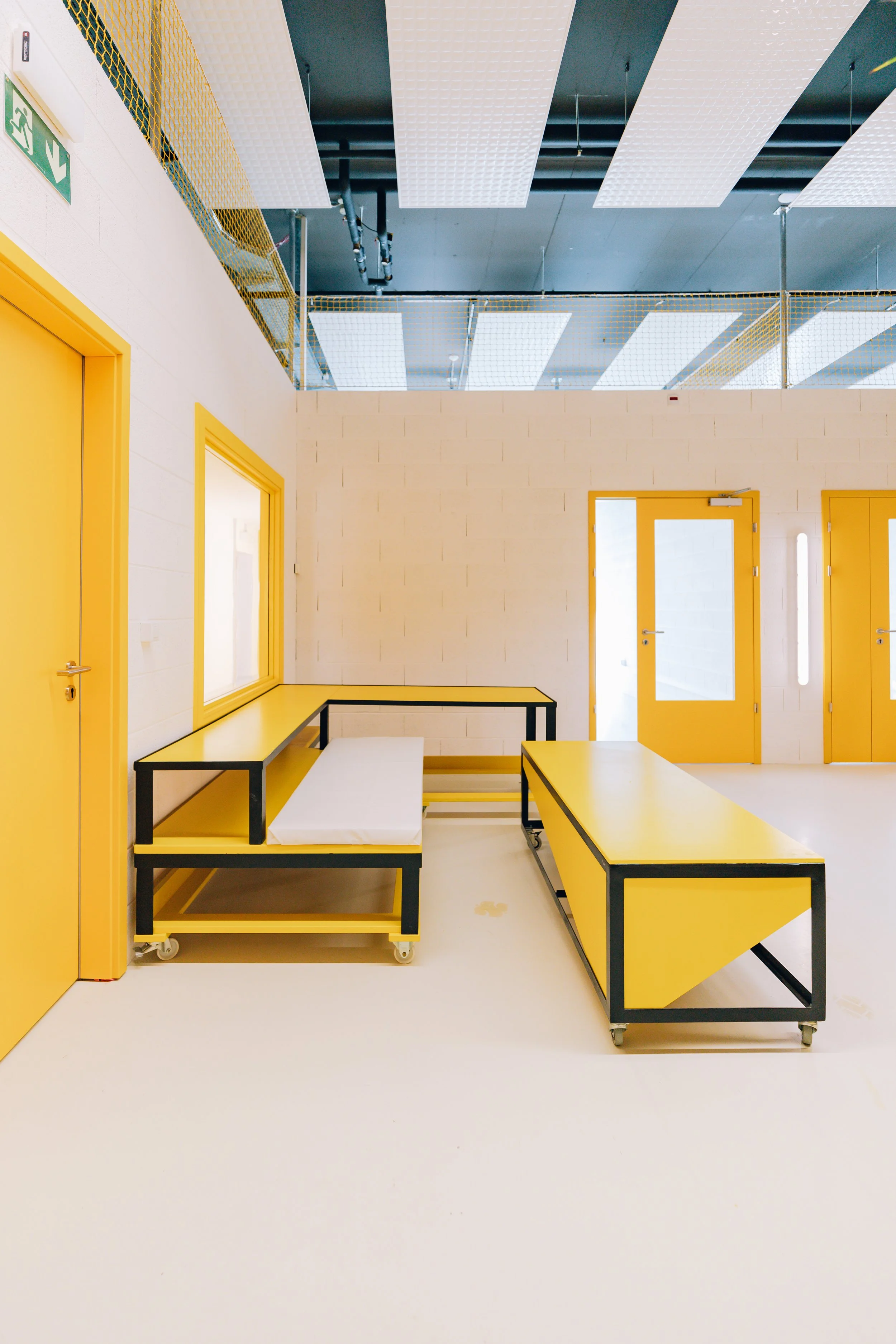 Intérieur d'une pièce avec murs blancs, portes jaunes avec vitrines, mobilier jaune et noir, plafonds avec panneaux blancs et tuyaux visibles, sol blanc, sortie de secours visible à gauche.