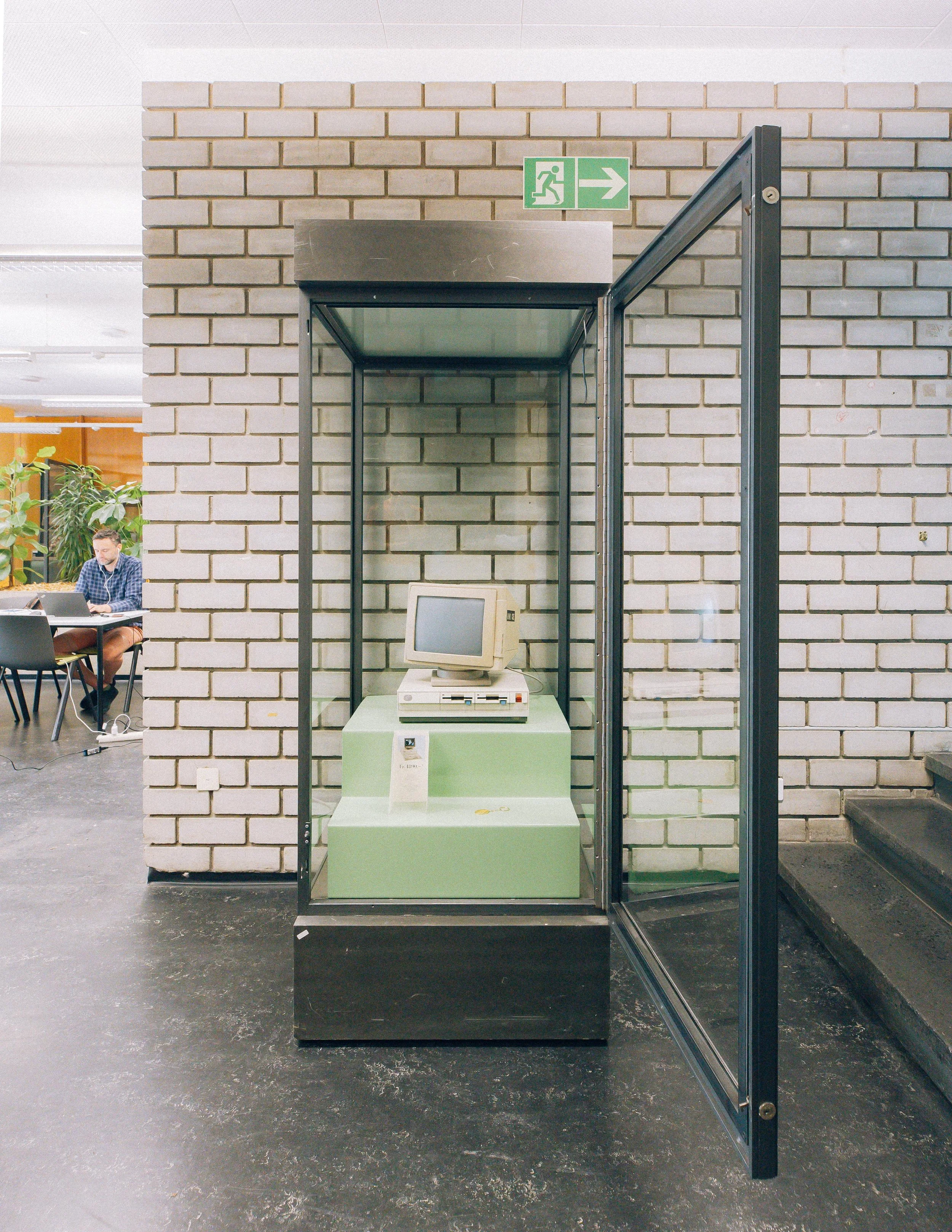Un étal d'exposition avec un vieil ordinateur de bureau en forme de monospace sur un socle vert. Derrière, un mur en briques et à gauche, une personne assise à une table utilise un ordinateur portable dans un environnement intérieur.