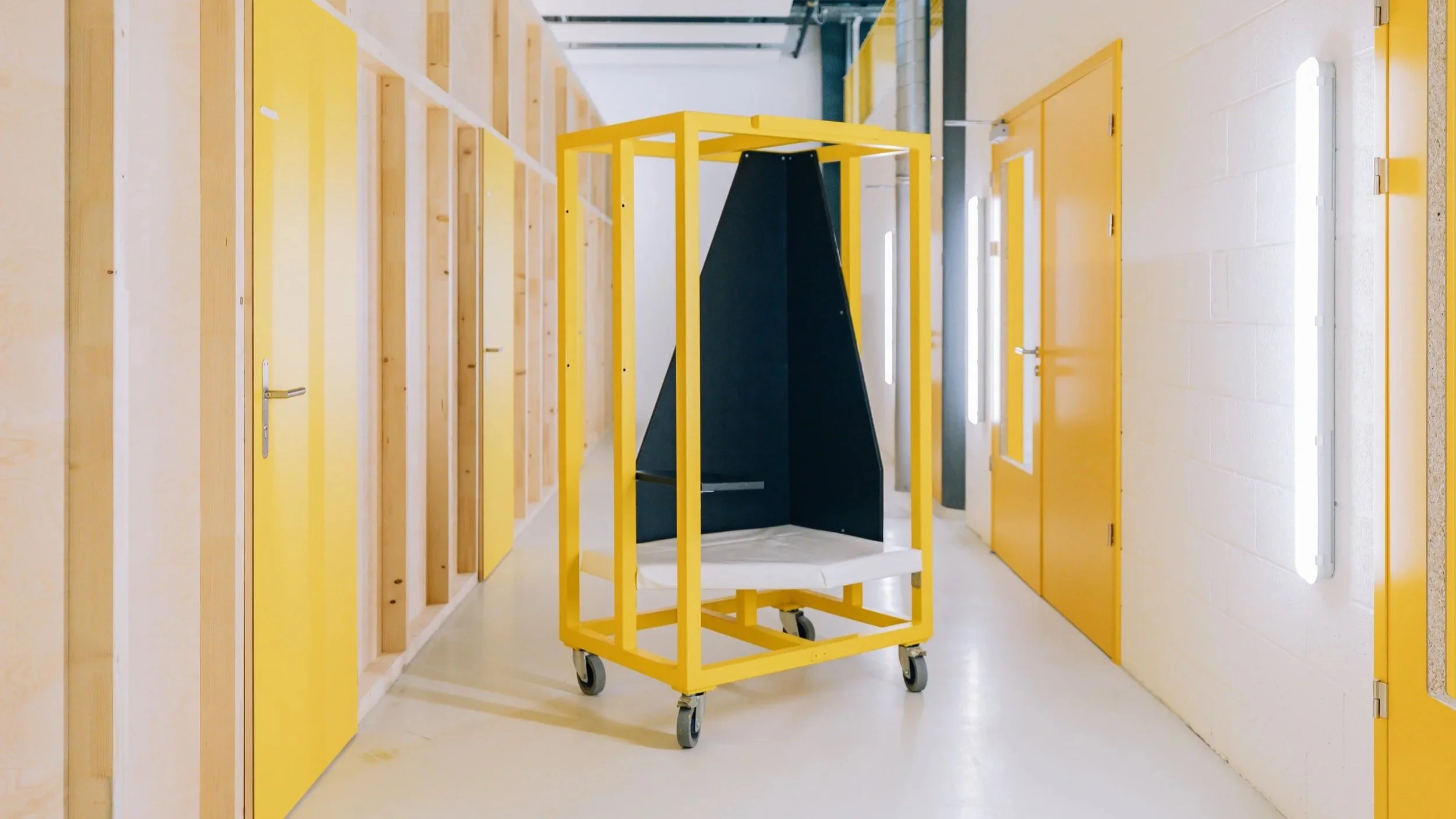 Alcôve téléphonique sur roulette, avec une assise en mousse de réemploi et bâche, une tablette. L'alcôve est dans un espace de coworking, avec des cloisons en bois, des portes jaunes et des néons blancs dans un espace industriel.
