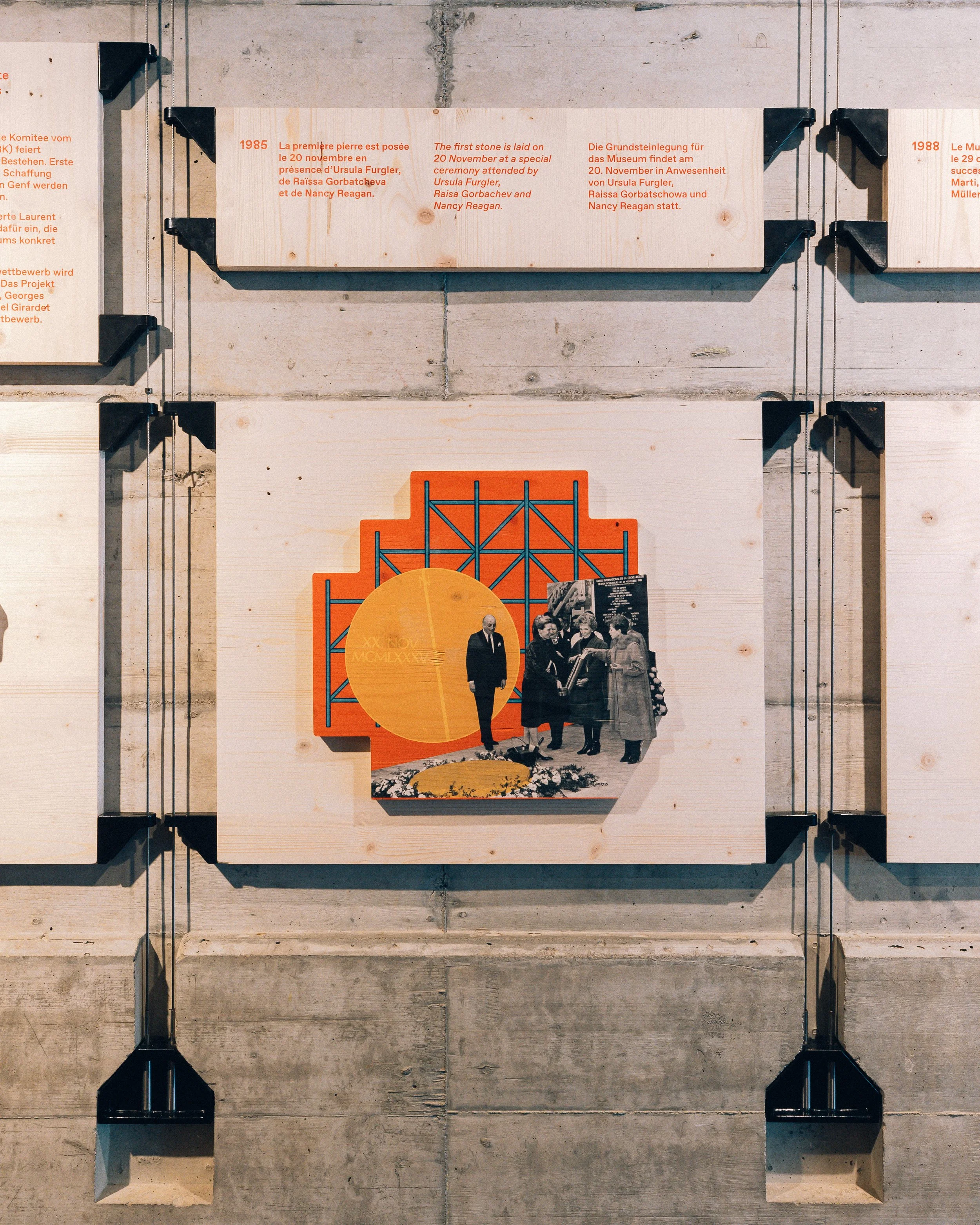 Affichage avec textes en français, anglais et allemand décrivant la pose de la première pierre lors d'une cérémonie en 1985, en présence d'Ursula Furgler, Raissa Gorbacheva et Nancy Reagan. Des éléments graphiques et une photo noir et blanc de plusieurs personnes en train de discuter figurent également en dessous.