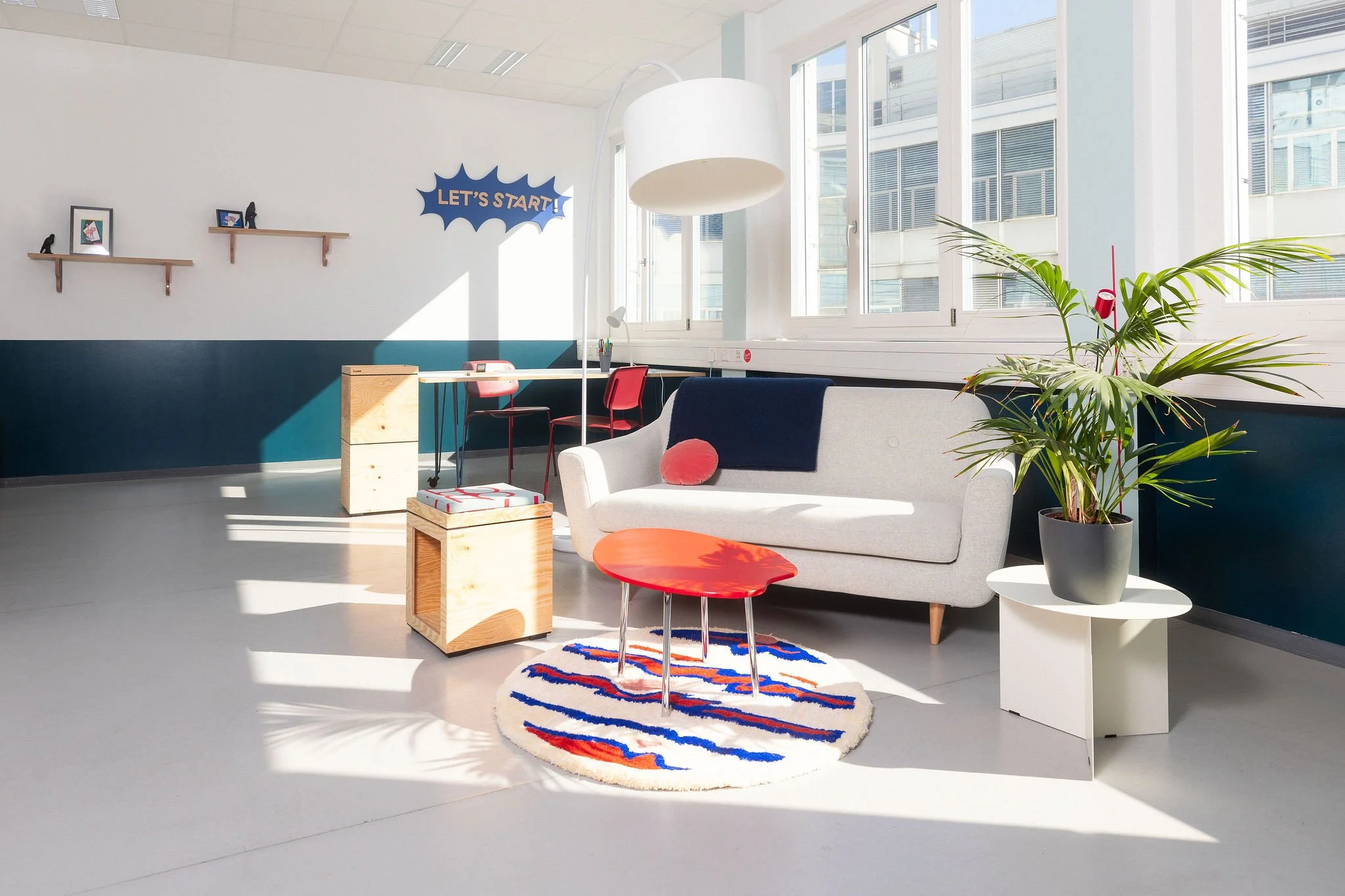 Bureau partagé dans coworking avec canapé, tapis rond à rayures rouges, blancs et bleues, meubles en bois clair, grande plante, murs blancs avec étagères en bois, fenêtre large et lumière naturelle, panneau "Let's start!" sur le mur.