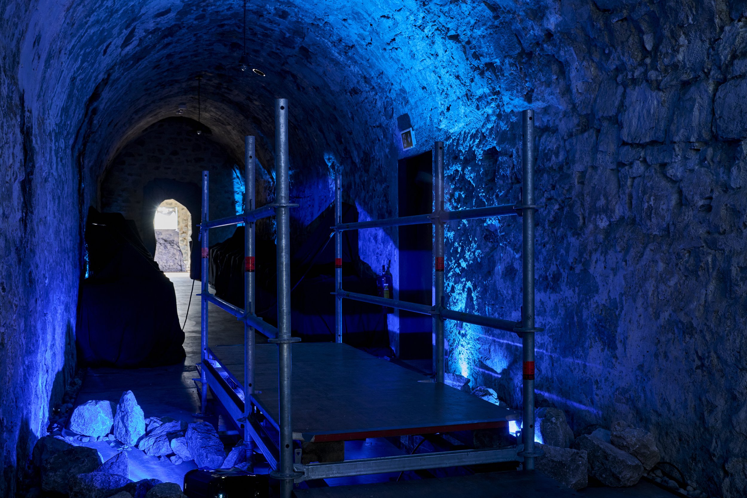 Intérieur d'une grotte avec des murs en pierre éclairés en bleu, présence d'une échelle en métal en cours de montage, et un homme casqué en silhouette.